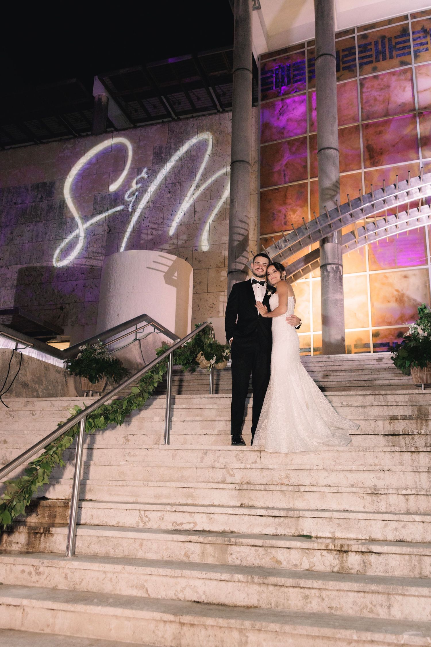 A wedding couple stands on elegant stone steps with illuminated purple decor and a glowing monogram on the wall.