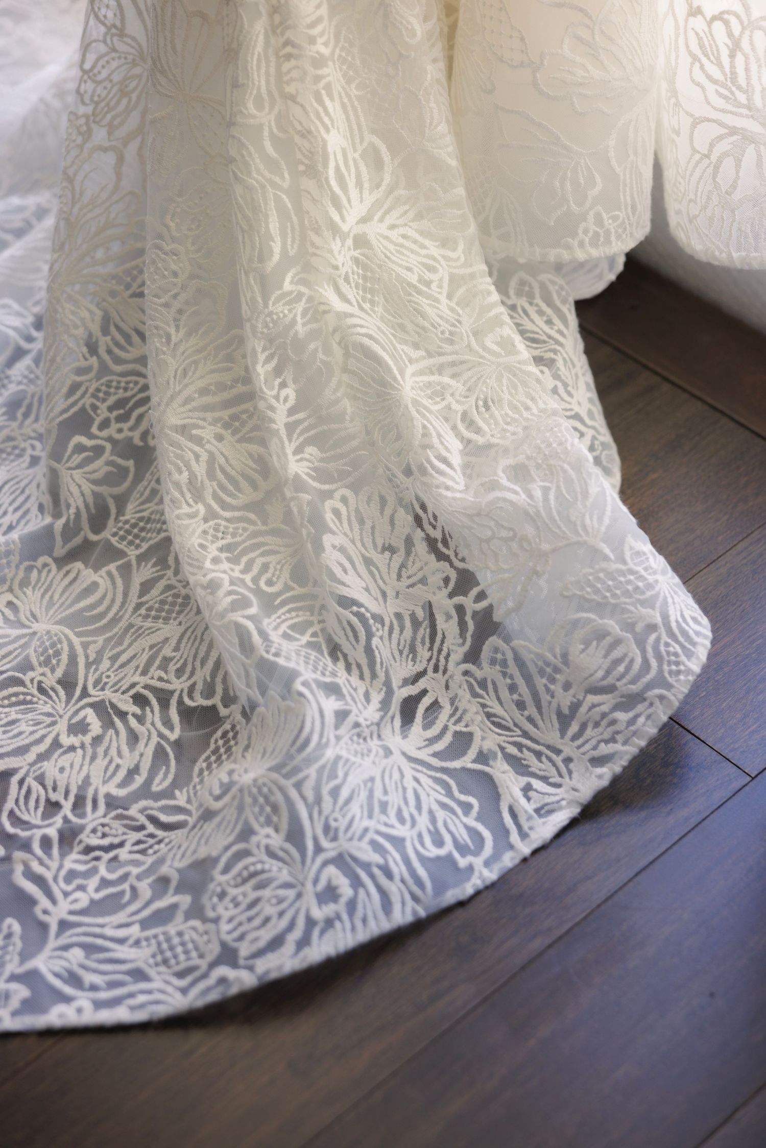 Close-up detail of delicate white lace wedding dress pattern against dark wooden floor.