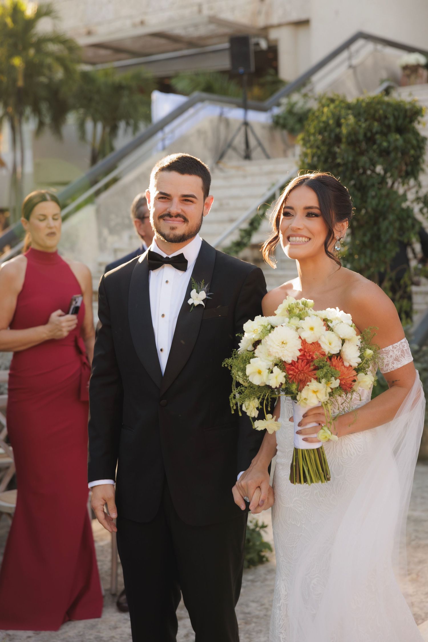 A wedding couple walks outdoors holding hands on their special day, dressed in formal black tuxedo and white gown.