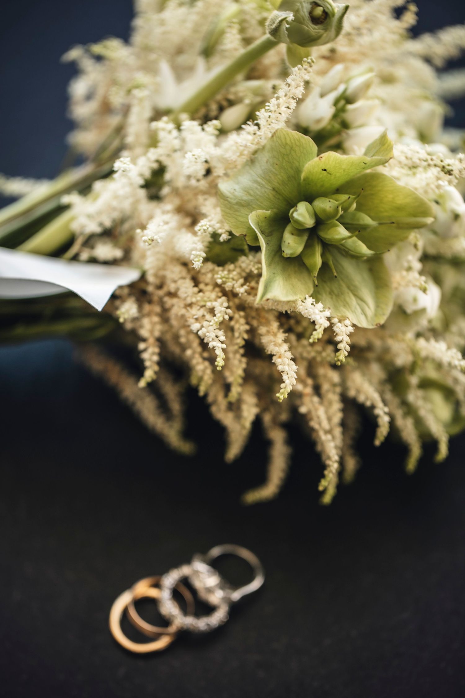 Delicate white floral bouquet with small blossoms and wedding rings against dark background.