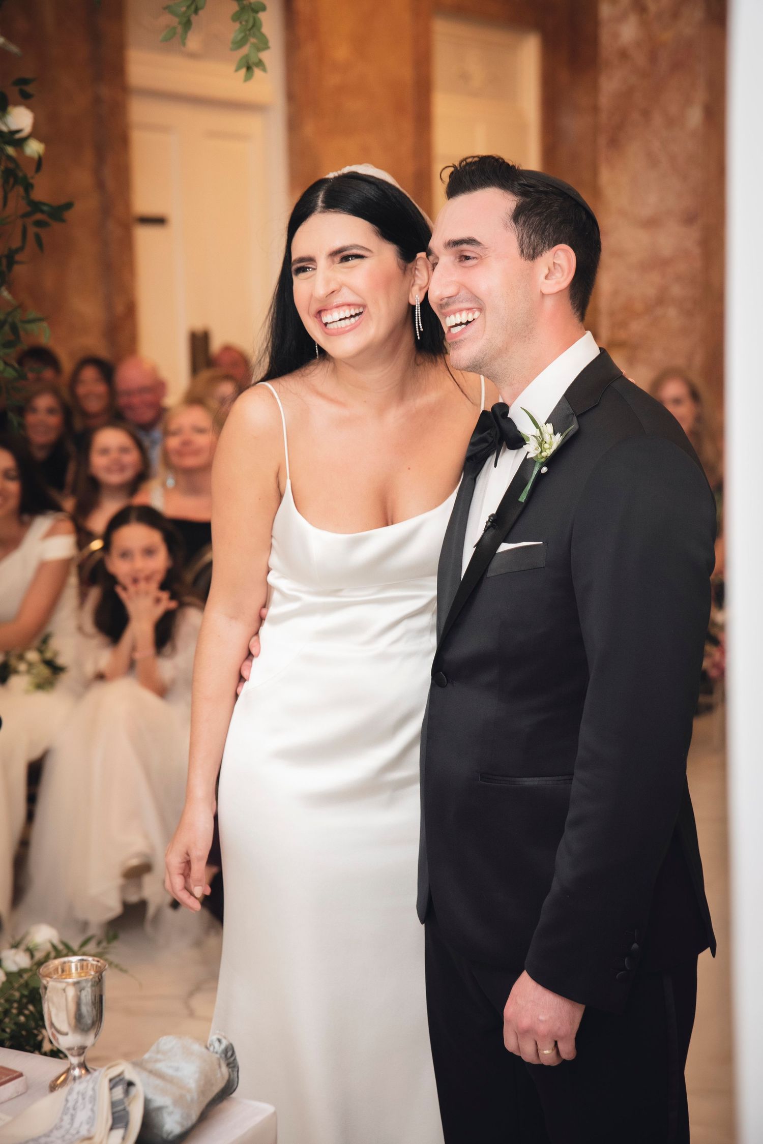 A happy couple share a joyous moment during their wedding reception in a warmly lit room with guests in the background.