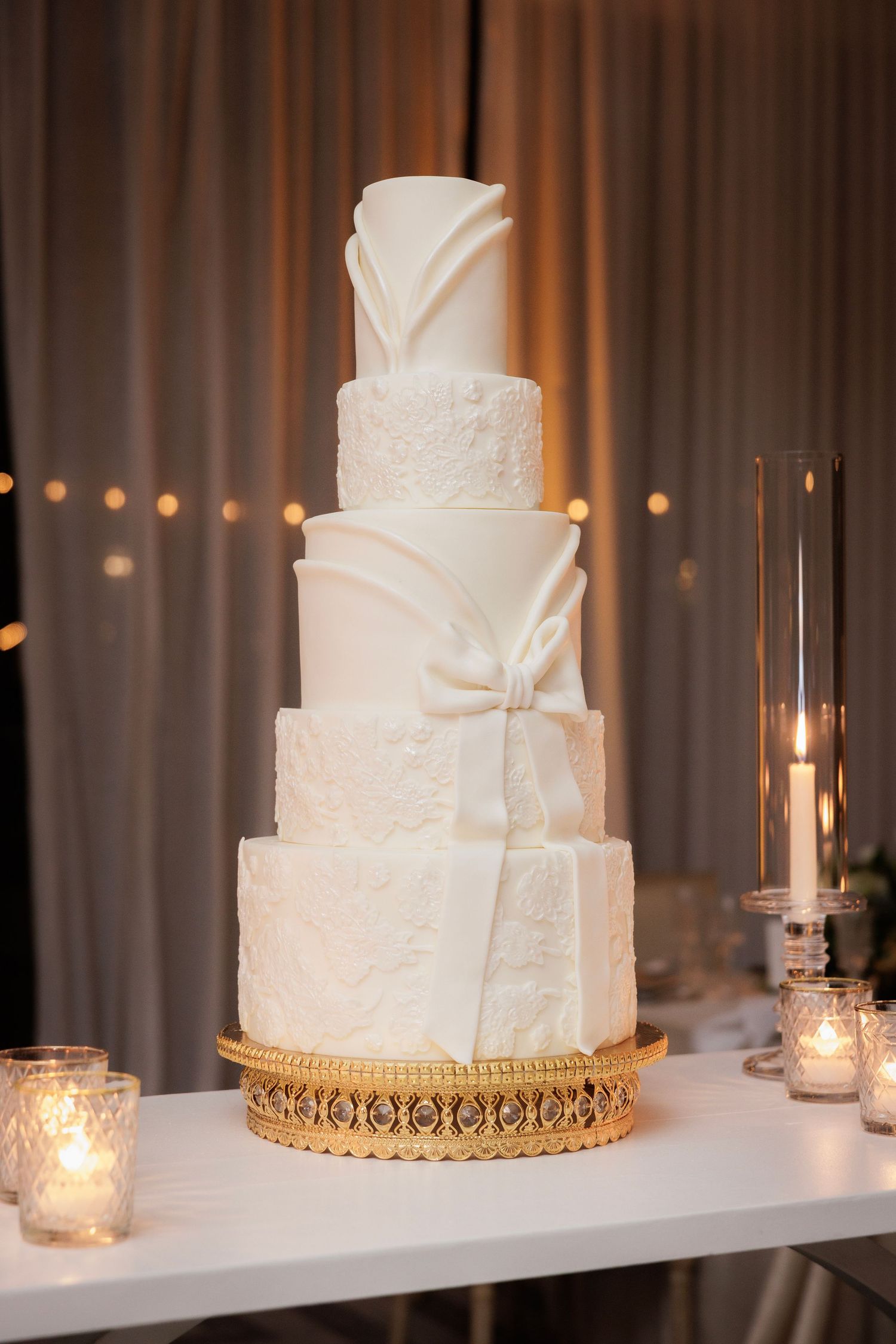 Three elegant white wedding cakes displayed on a white shelf with soft candlelight and gold accents create a romantic atmosphere.