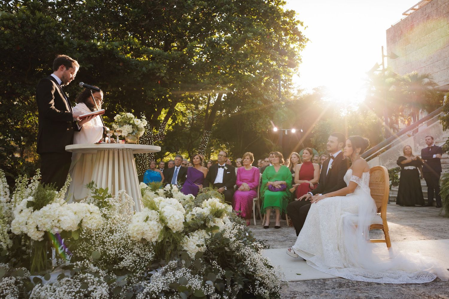 An outdoor wedding ceremony at sunset with floral arrangements, guests seated on chairs, and a couple at an altar.