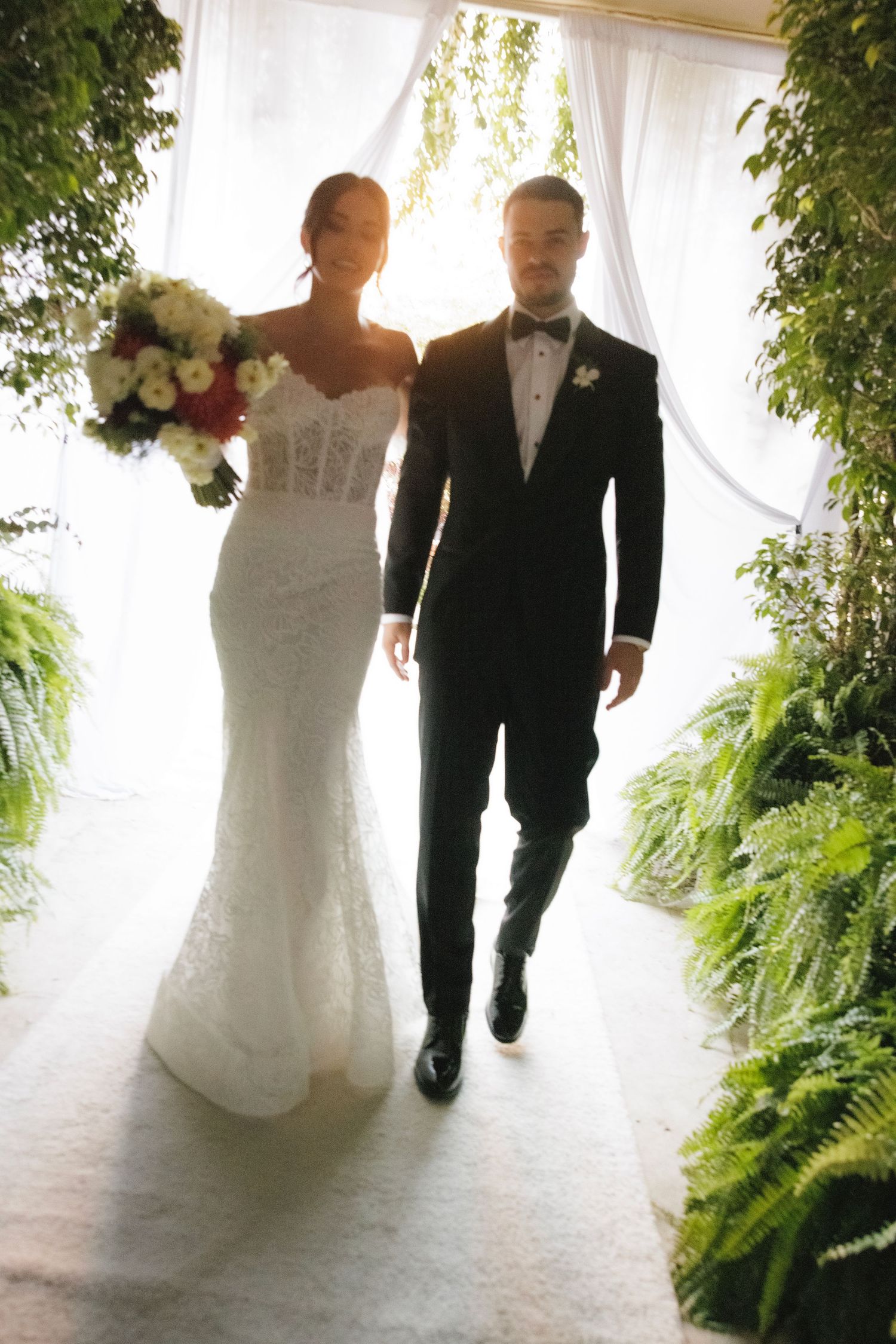 A wedding couple walks through an elegant white-draped corridor lined with potted ferns creating a romantic ambiance.