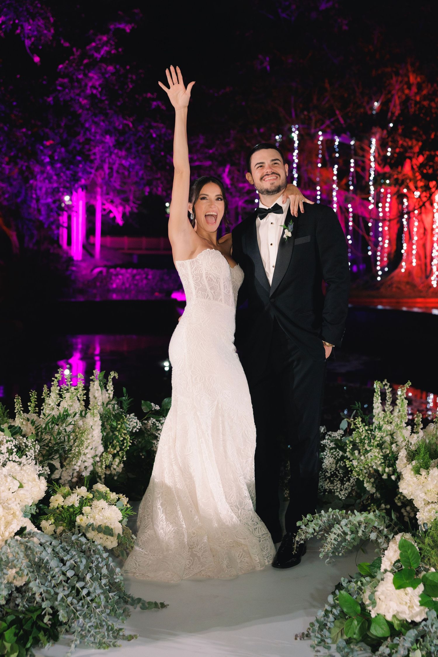 Newlyweds celebrate under purple and red outdoor lighting with floral decorations and string lights.