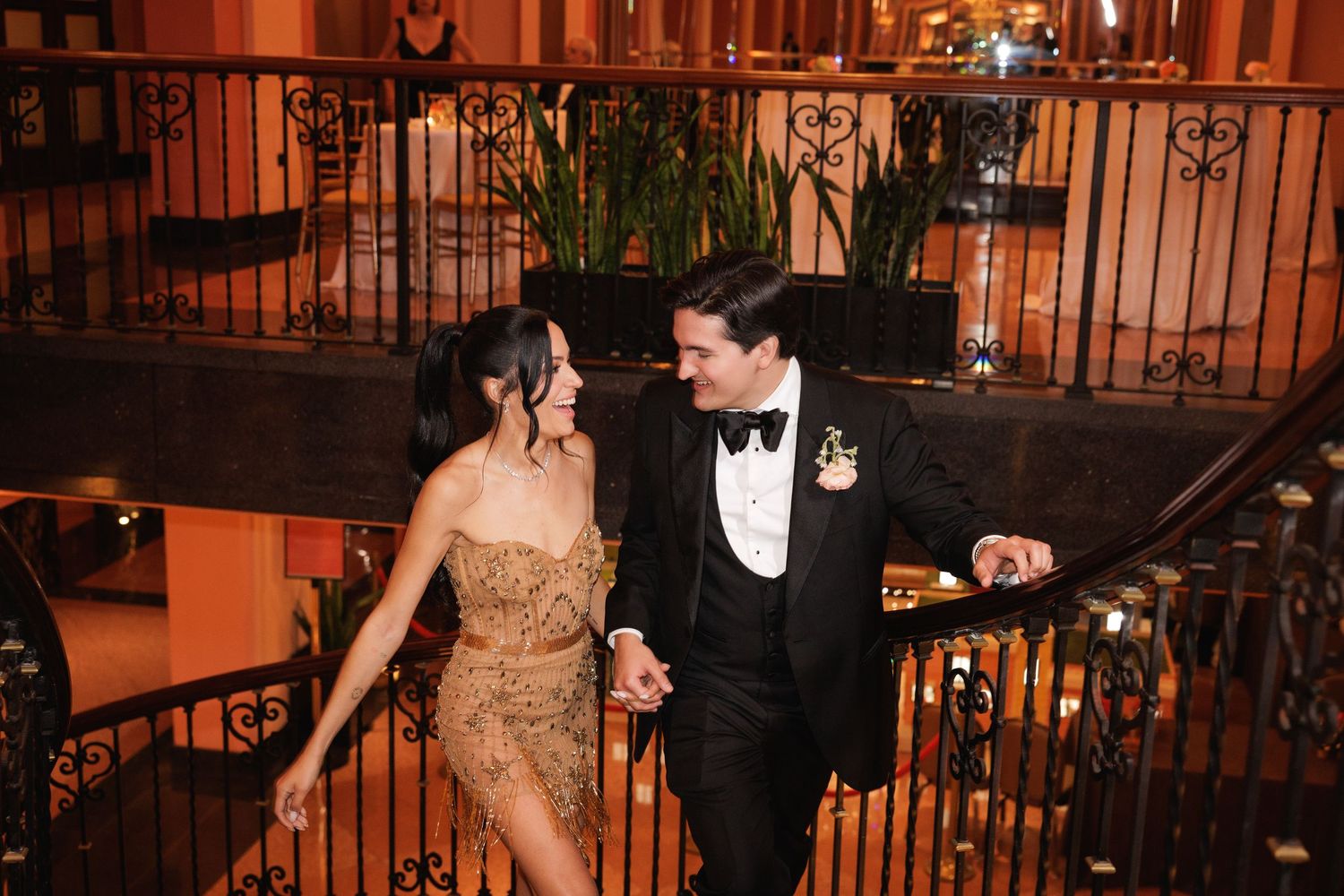 A couple in formal attire descend an ornate staircase at an elegant venue illuminated in warm amber lighting.