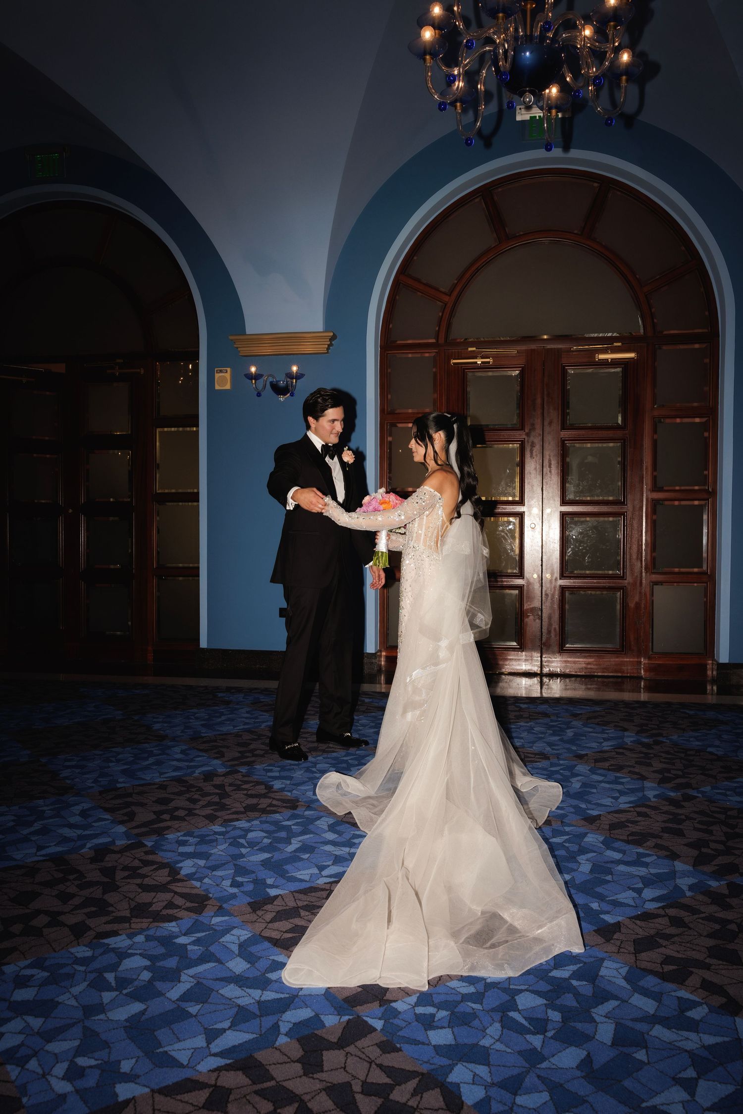 Newlyweds sharing their first dance in a grand ballroom with ornate wooden doors and blue arched ceilings.