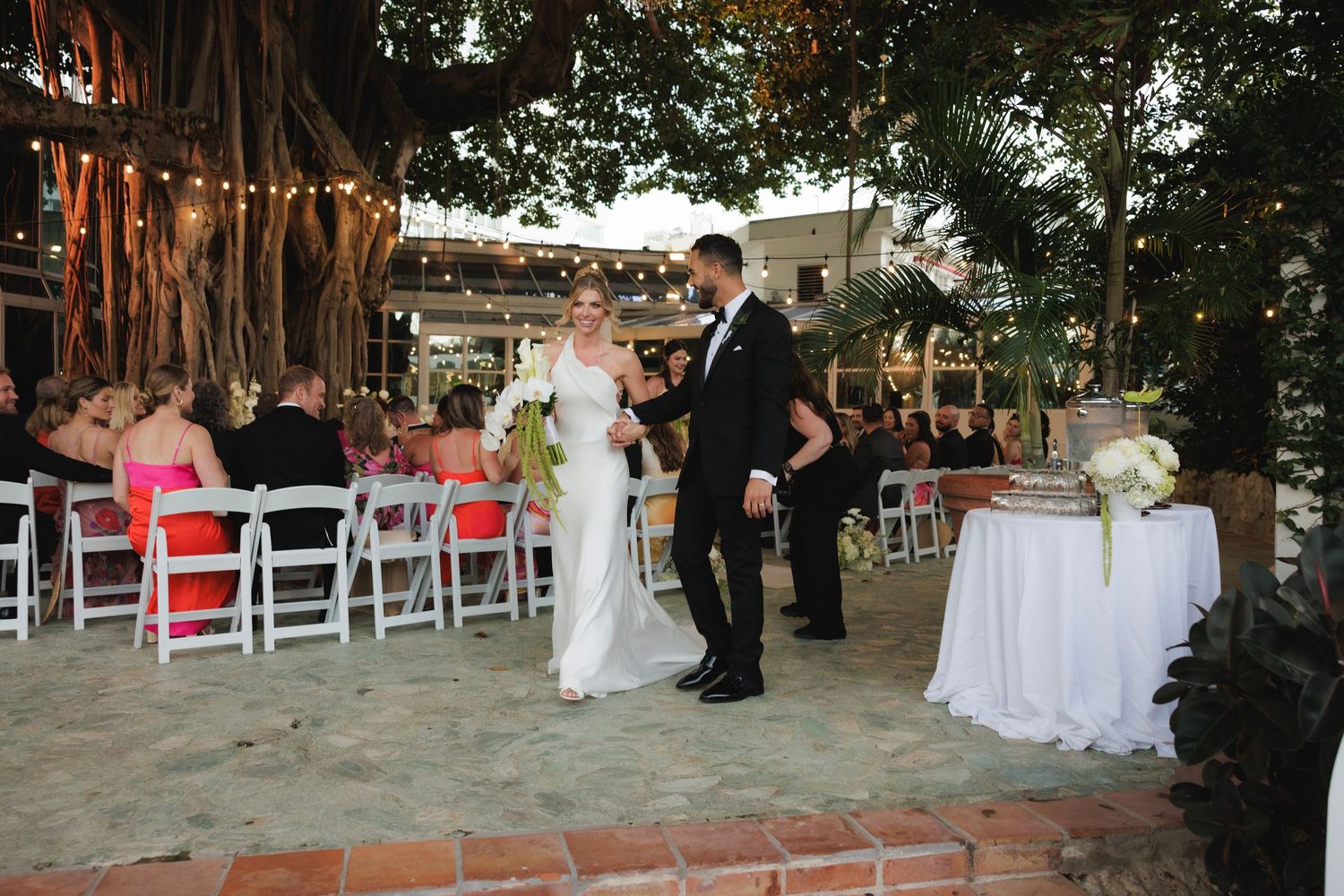 Wedding ceremony takes place outdoors under overhanging tree branches with white chairs and decorations.