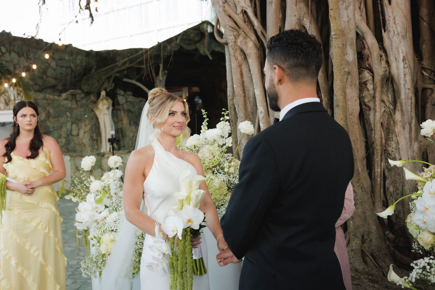 Wedding ceremony takes place beneath rustic wooden arch with white floral decorations and string lights.