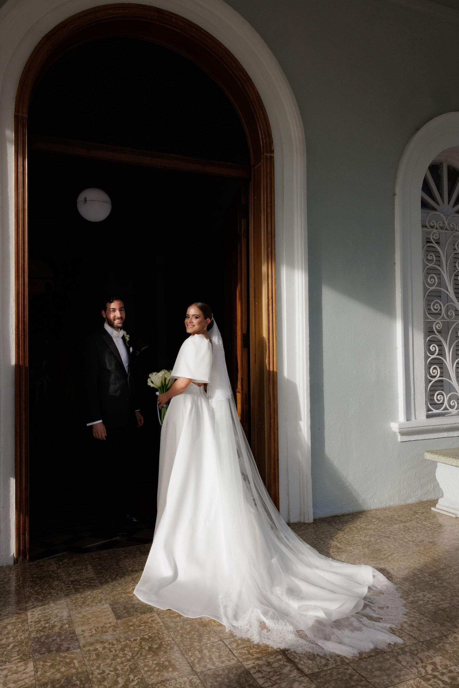 A wedding couple poses in an arched doorway casting dramatic shadows on a white stone facade.