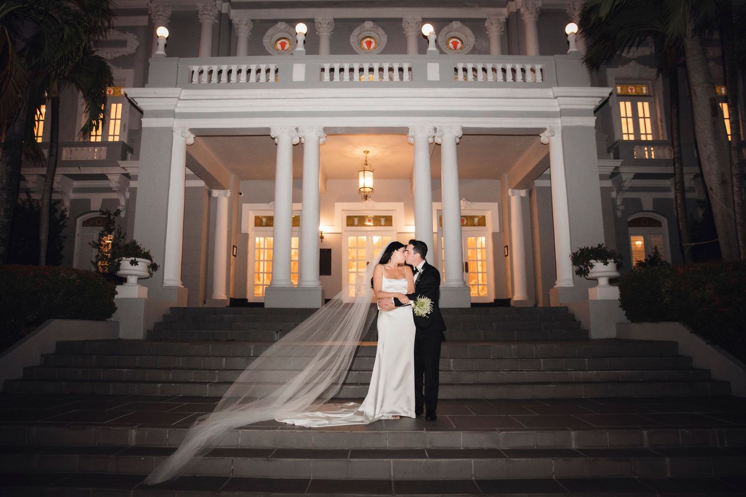 A romantic nighttime portrait of newlyweds standing on the steps of an elegant white columned mansion with glowing lights.