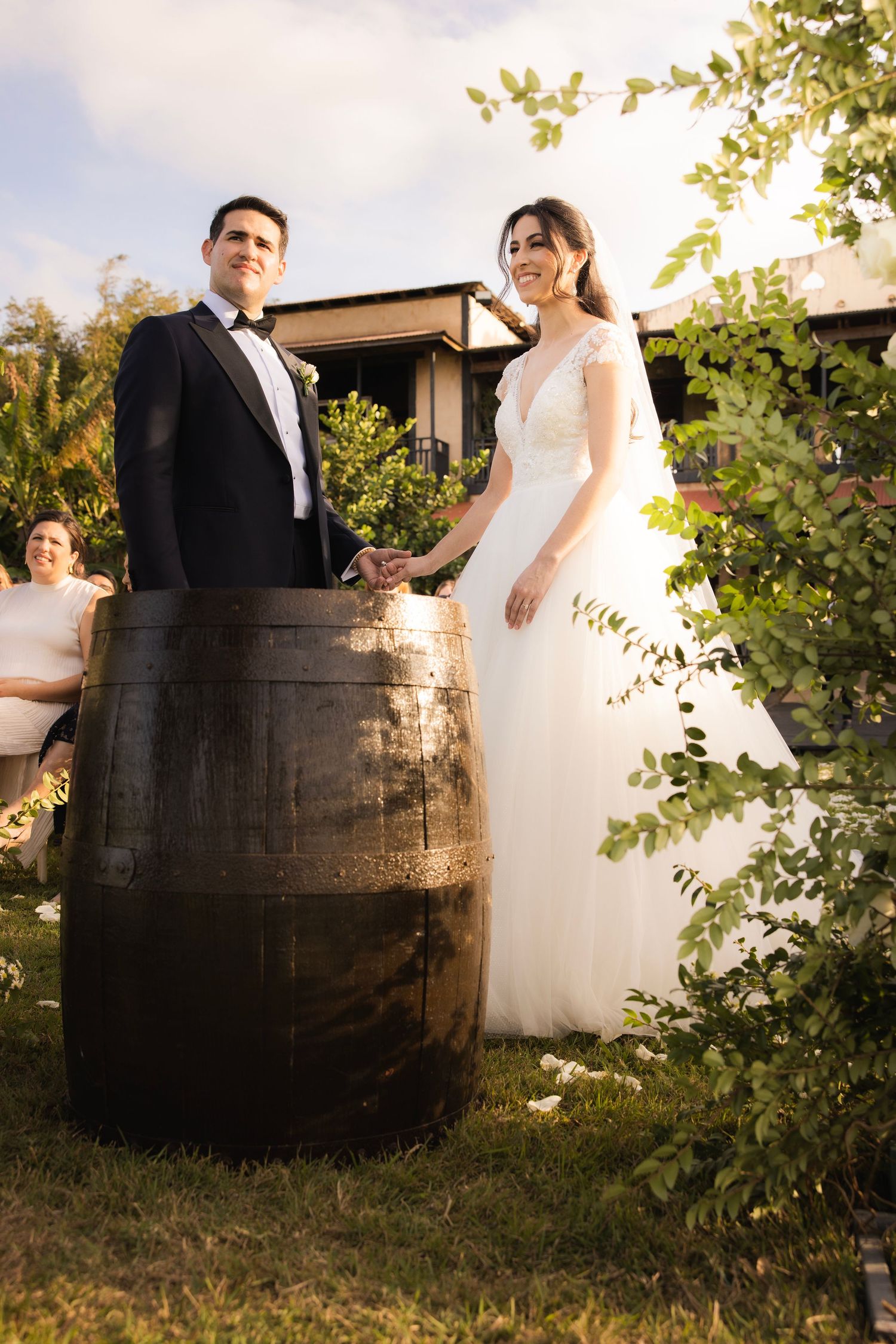 Newlyweds pose for rustic wedding photos with wooden wine barrels in a beautiful garden setting.