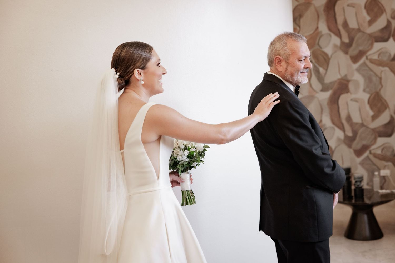 A bride and groom share an emotional first look moment in a softly lit hallway with neutral tones and textured walls.