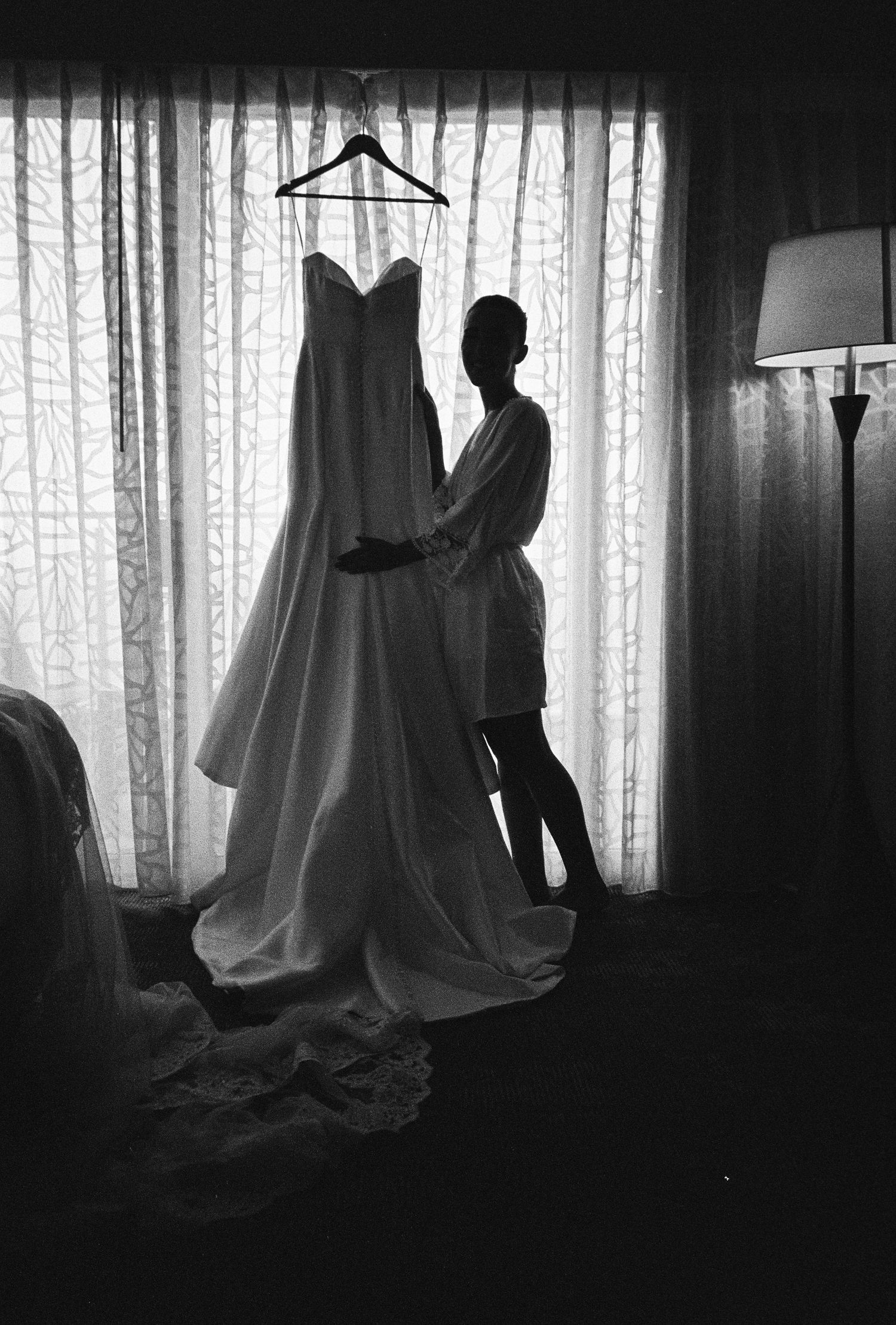 Silhouette of a bride holding her wedding dress on a hanger in front of a bright window.