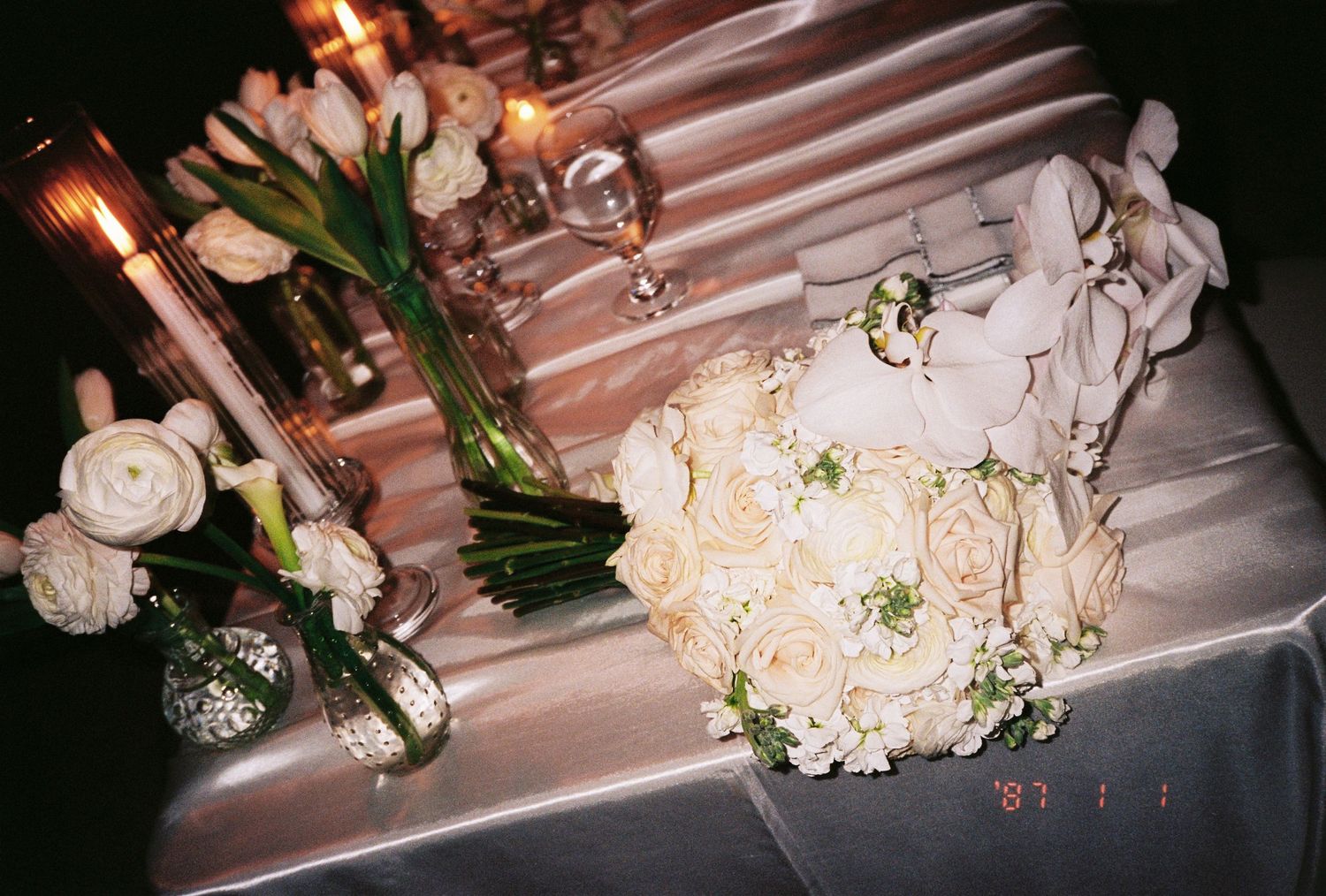 Elegant wedding table with white rose bridal bouquet, tulips, and candlelight centerpieces on a satin tablecloth.