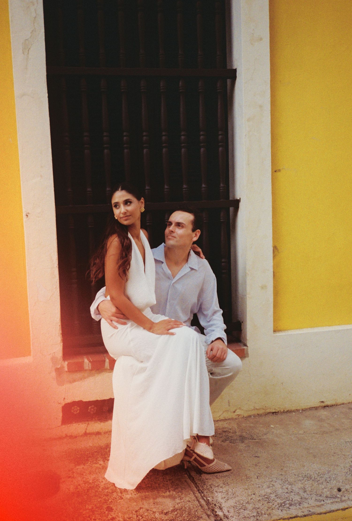 Couple posing in front of yellow building, woman in white dress sitting on man's knee in Old San Juan.