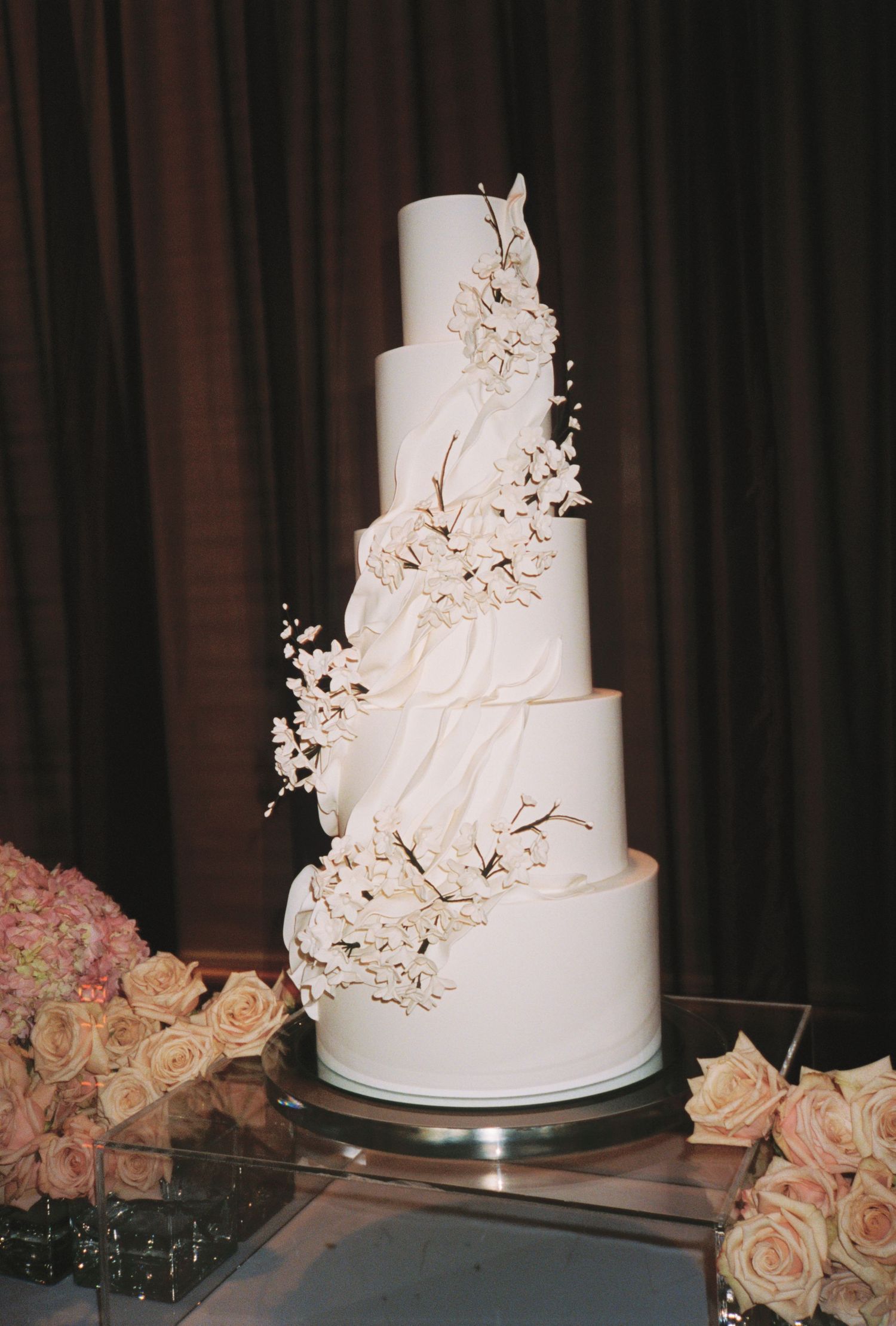 Elegant four-tier white wedding cake with cascading cherry blossom branches, displayed beside pink roses.