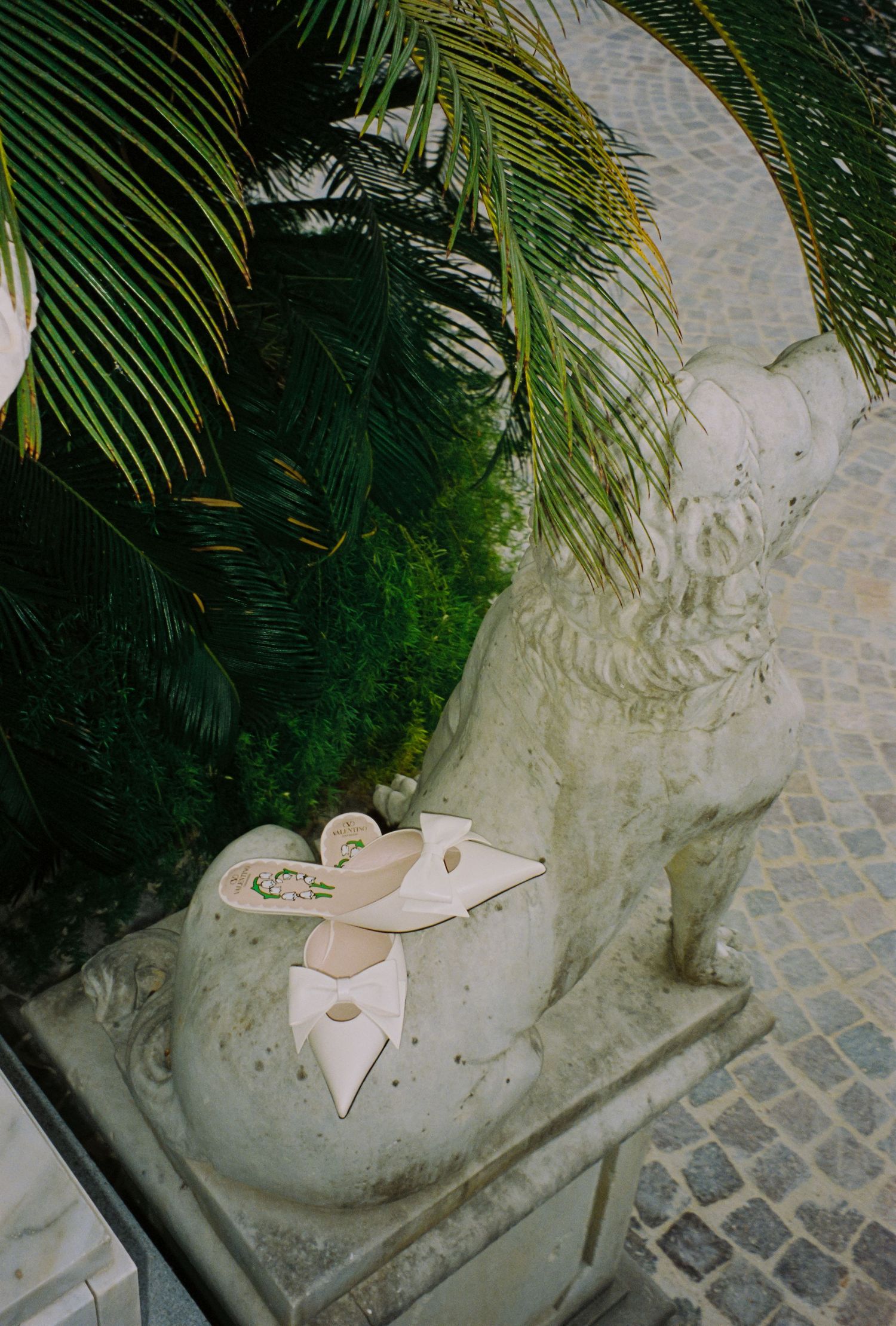 White heeled mules with floral detail rest on a stone lion sculpture surrounded by lush tropical greenery.