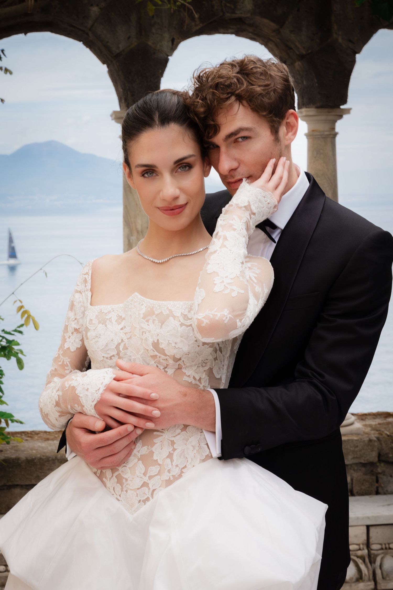 Elegant bride and groom embrace by lakeside archway, she in lace wedding gown, he in black tuxedo.