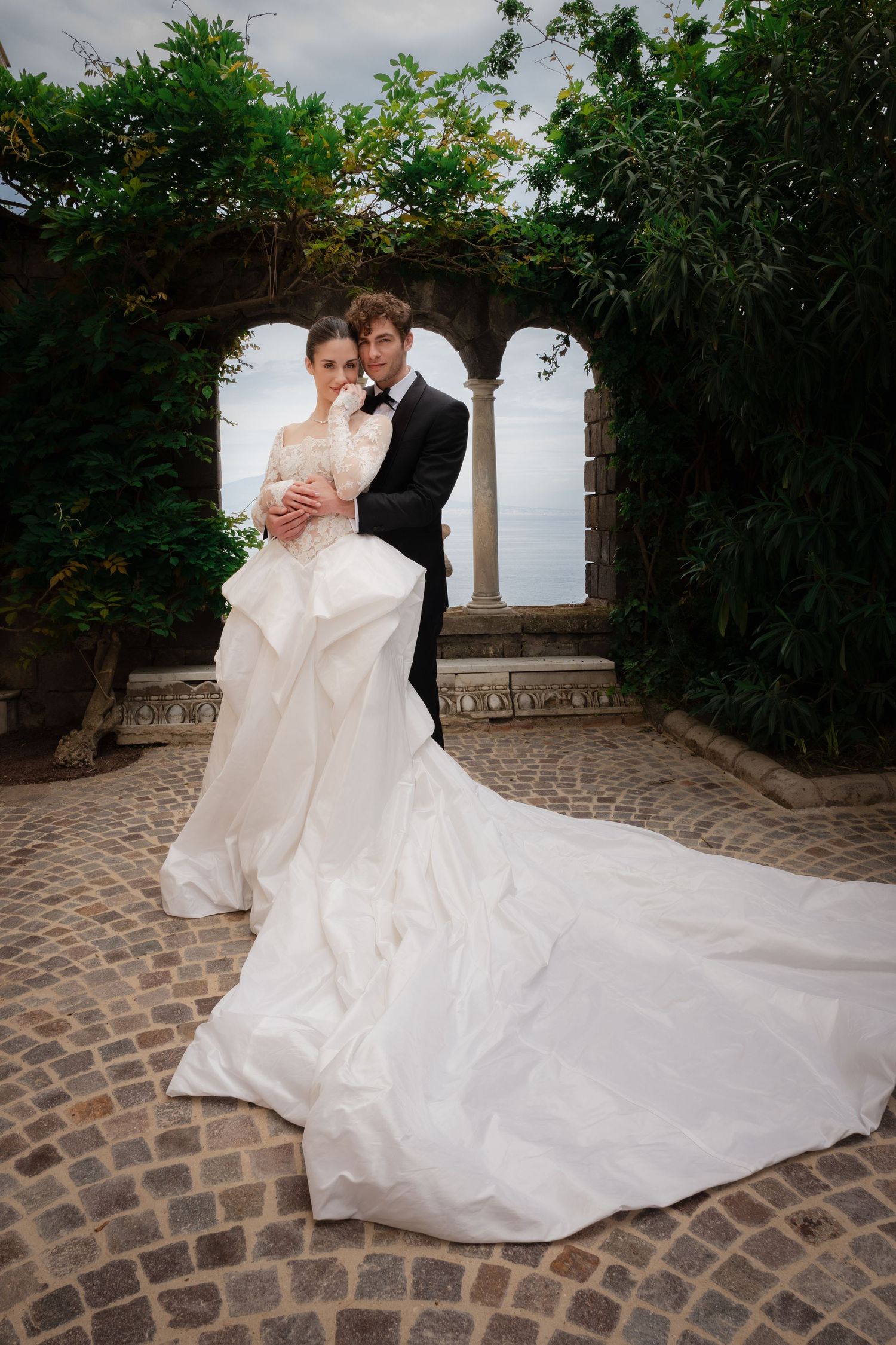 Bride in elegant white gown with long train embraces groom in tuxedo under stone arch with lush greenery.