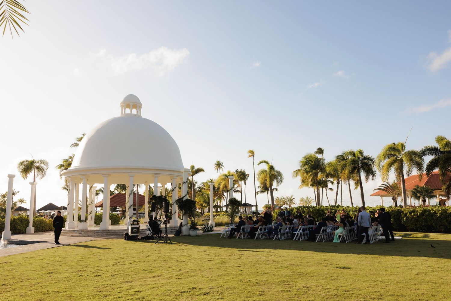Panoramic view of a beachside wedding ceremony with white gazebo, palm trees, and guests seated on chairs.