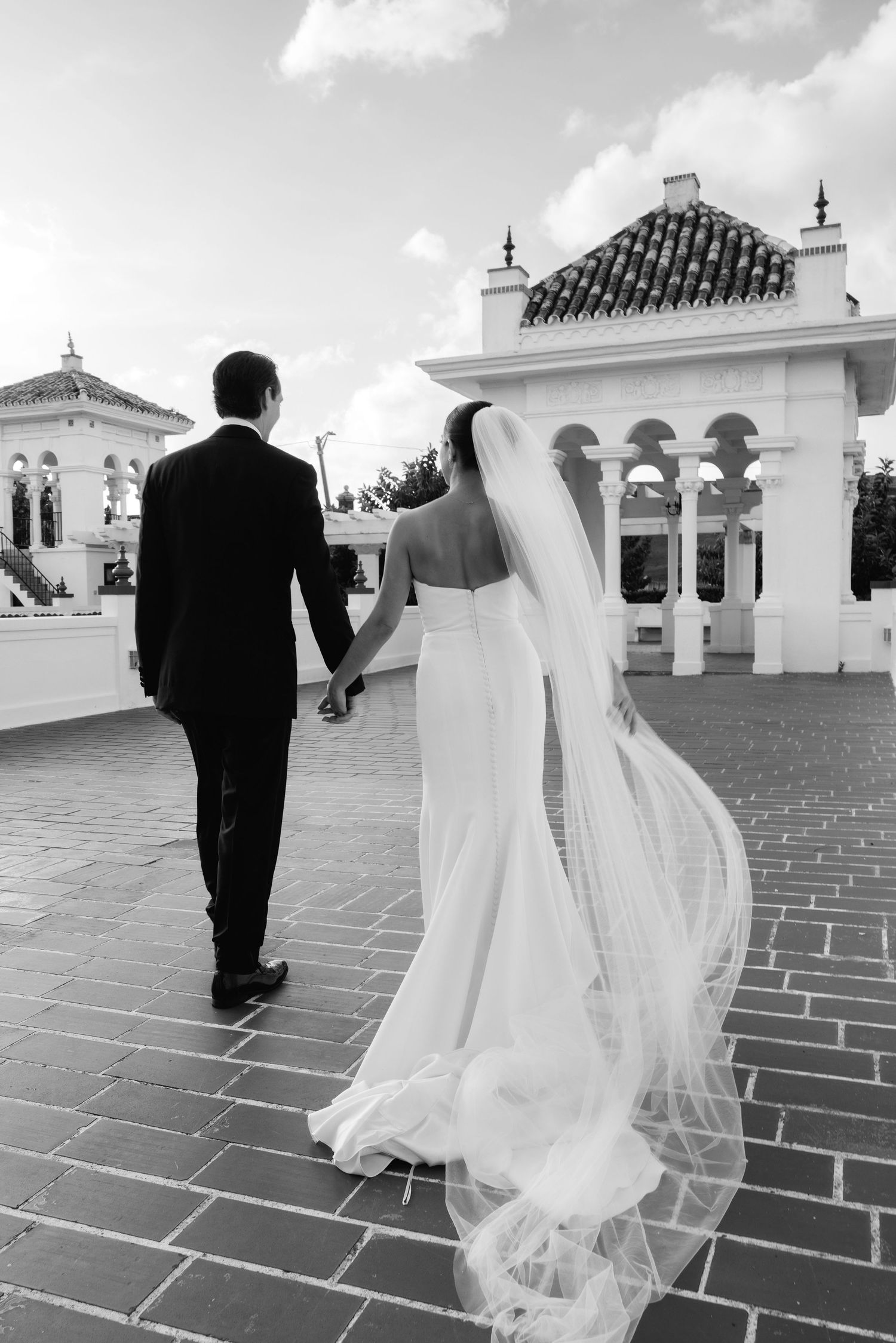 Bride and groom walk across Spanish-style rooftop terrace under bright sky.