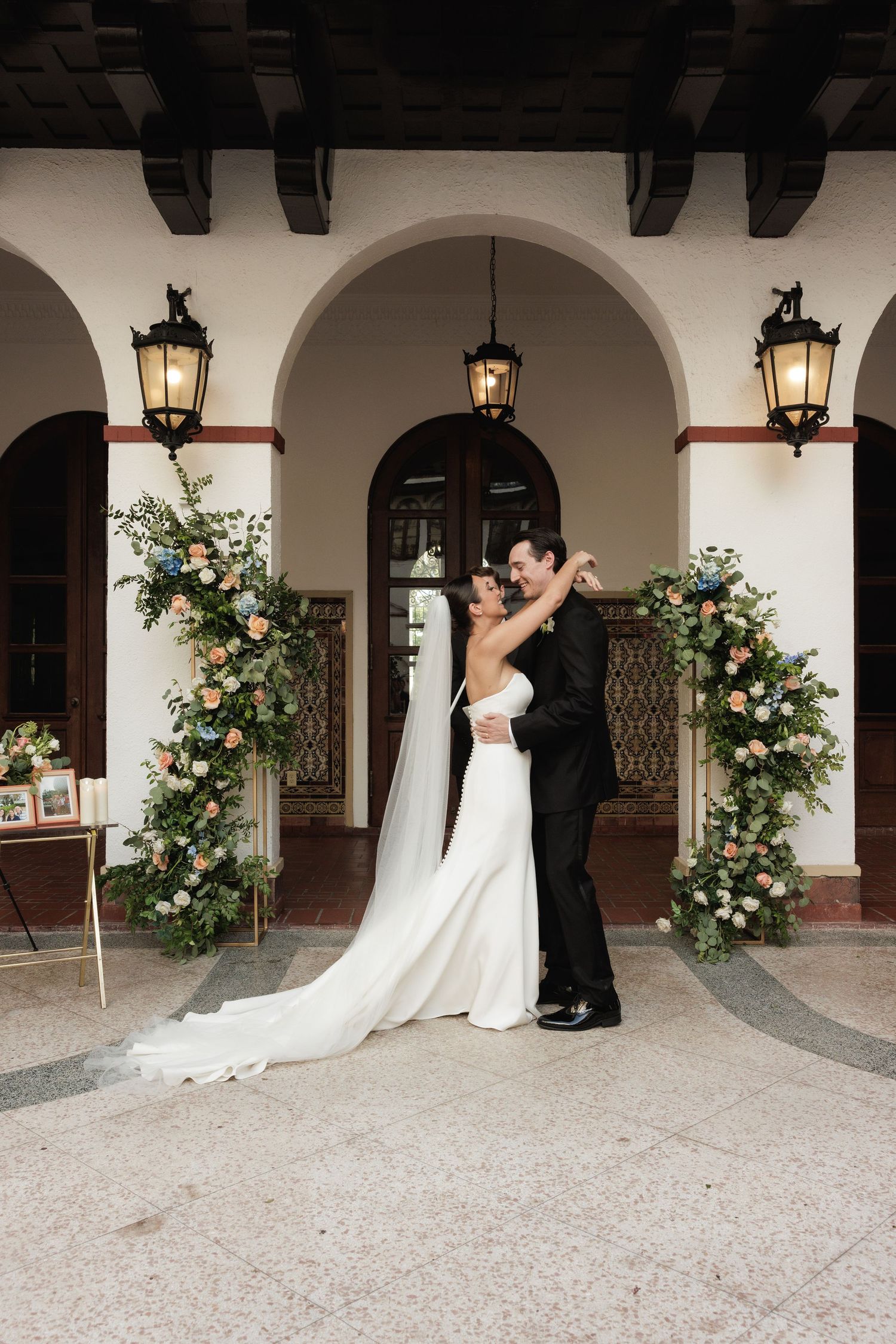 A wedding couple embraces under arched walkway with hanging lanterns and floral arrangements at elegant venue.