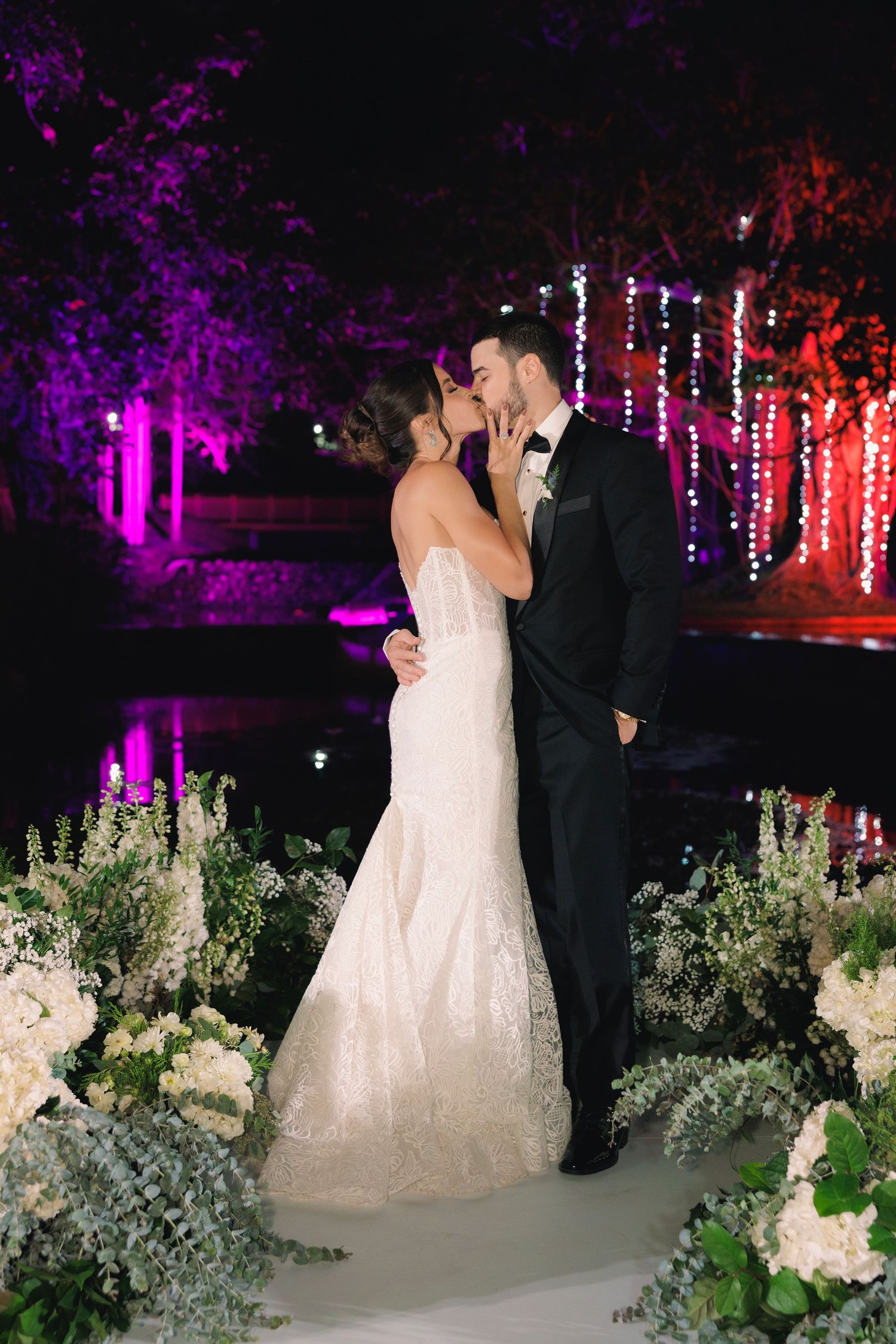 A romantic wedding scene at night with purple lighting and white floral arrangements decorating the outdoor venue.