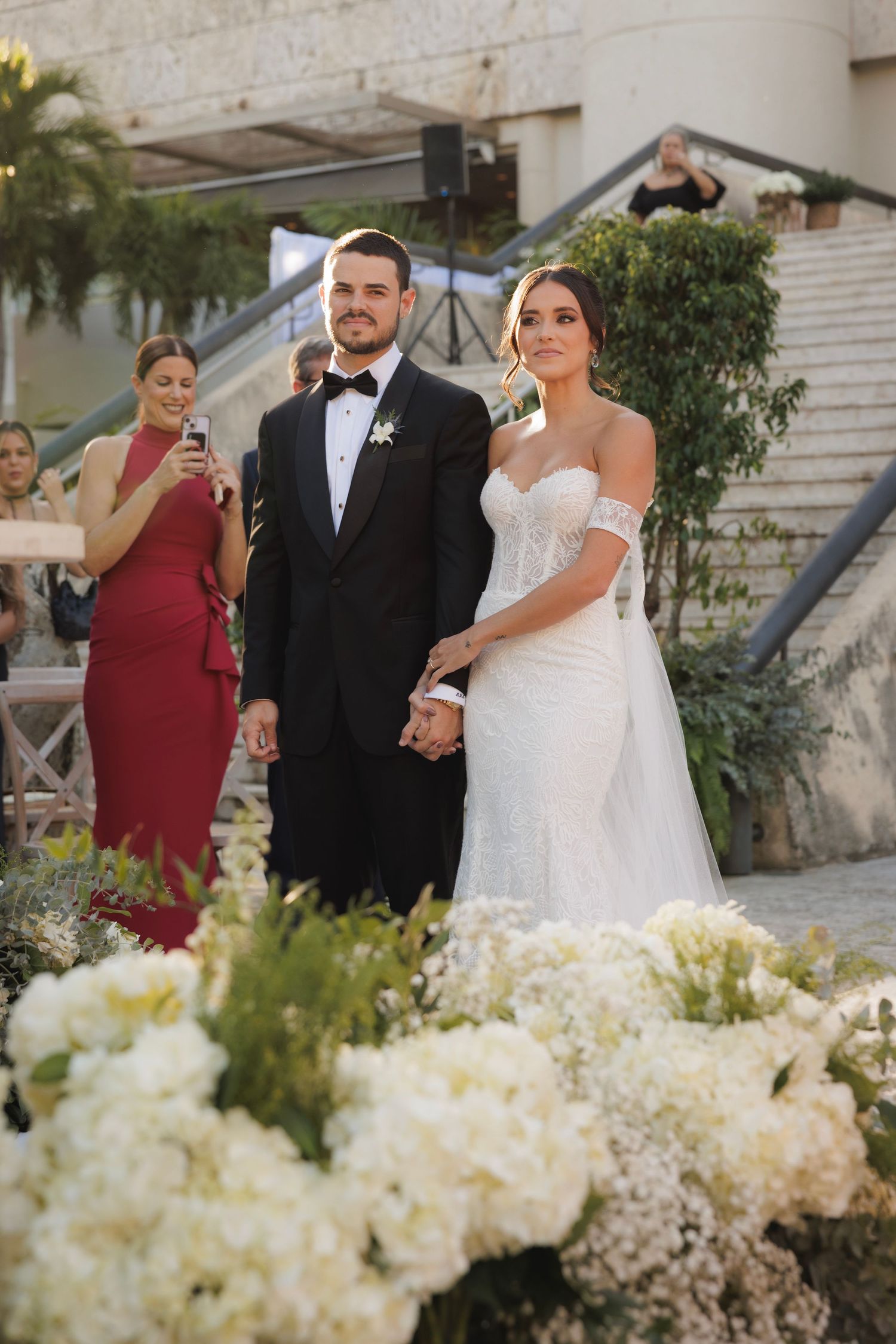 Wedding ceremony setup with white floral arrangements and guests gathering on stone steps.
