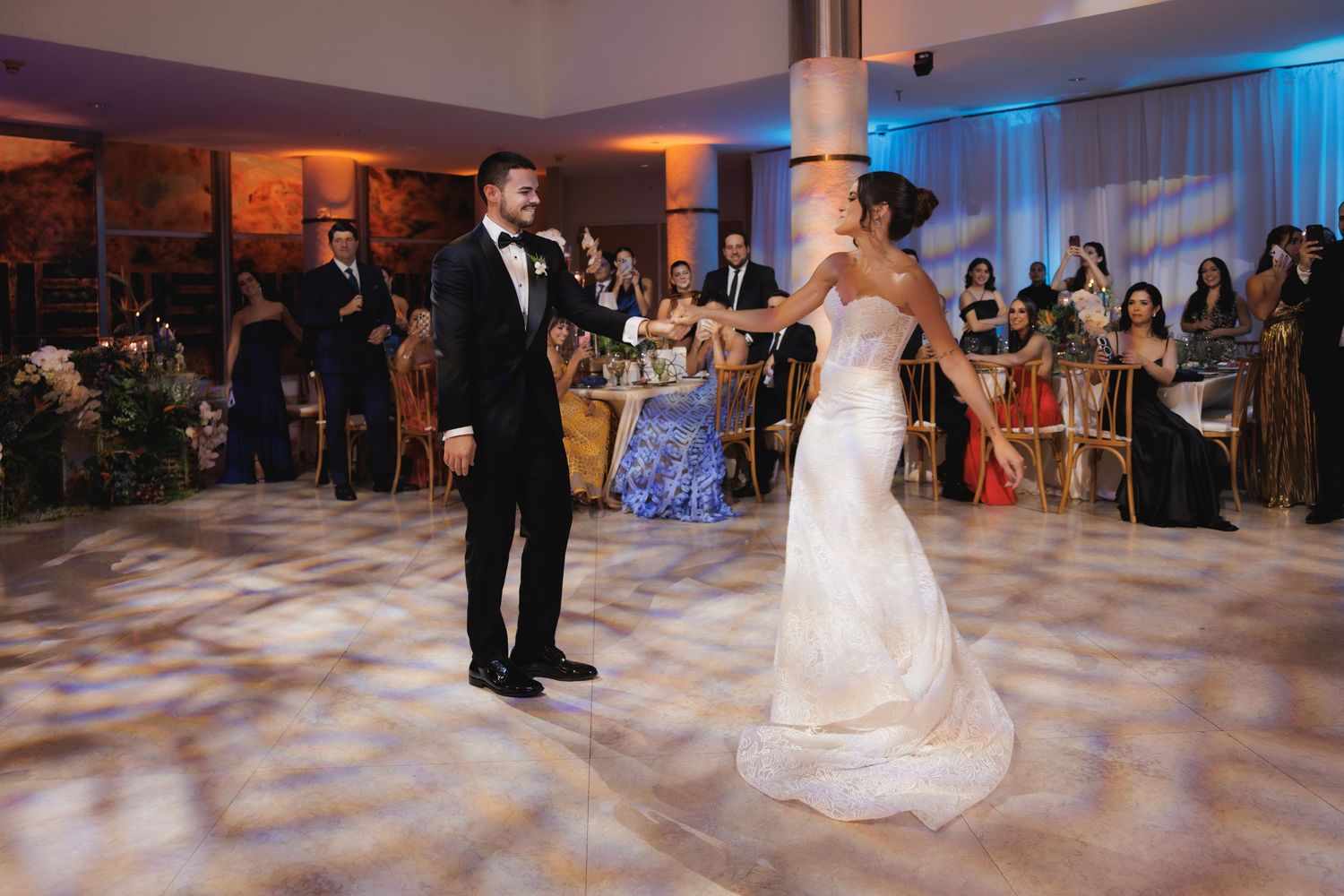 Newlyweds perform their first dance together on an illuminated ballroom floor surrounded by wedding guests.