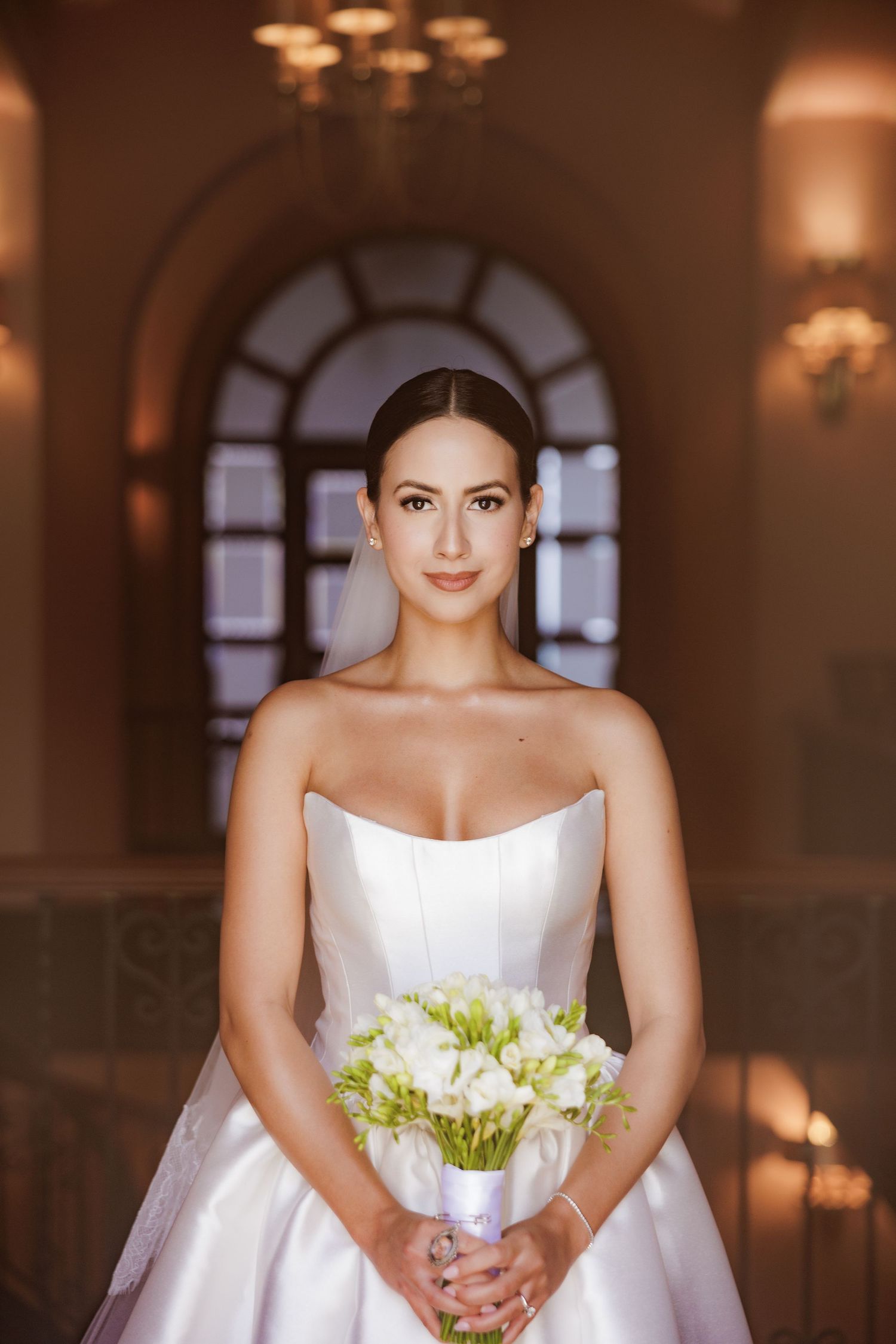 A bride in a white strapless wedding gown holds a bouquet of white and green flowers in a warmly lit venue.