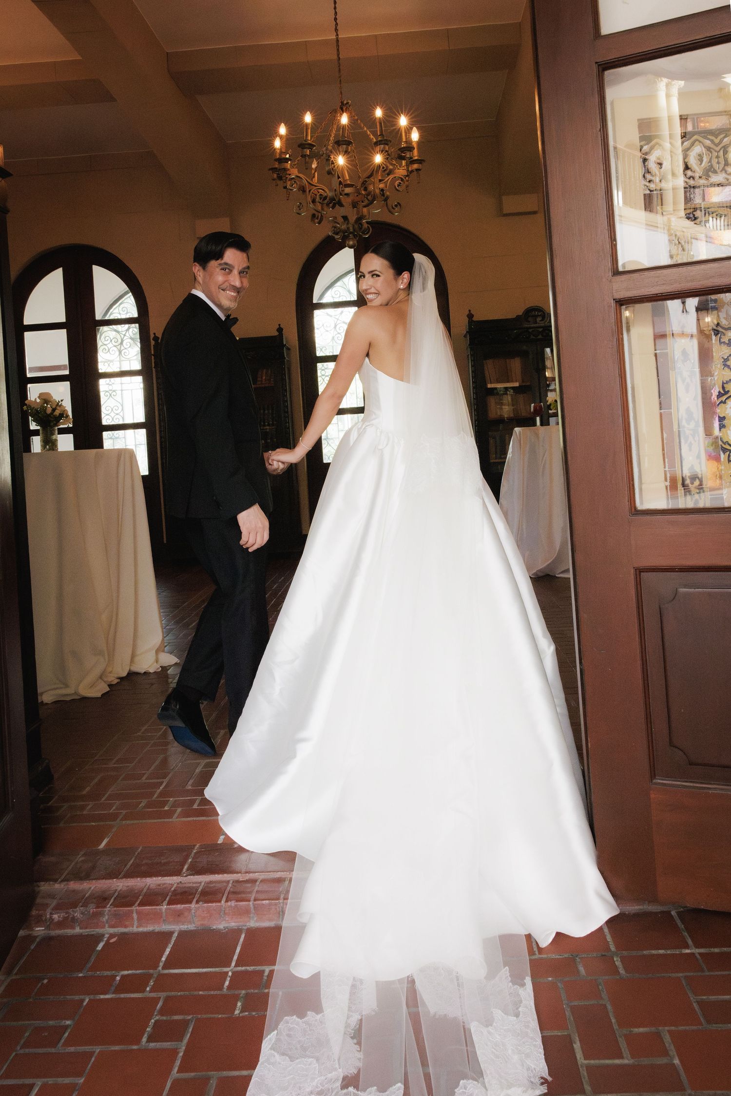 Elegant bride in flowing white gown walks across terracotta tile floor in historic venue with arched windows and chandelier.