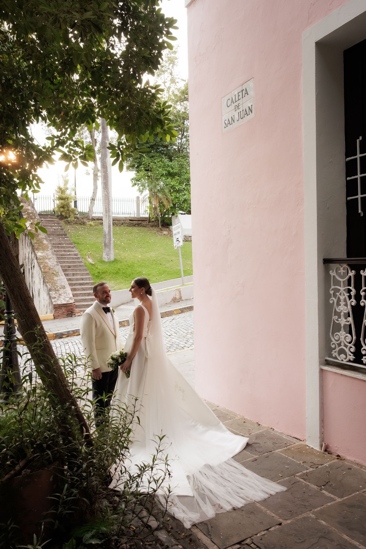 A wedding couple stands on a stone pathway next to a pink building with trailing white dress and lush greenery overhead.