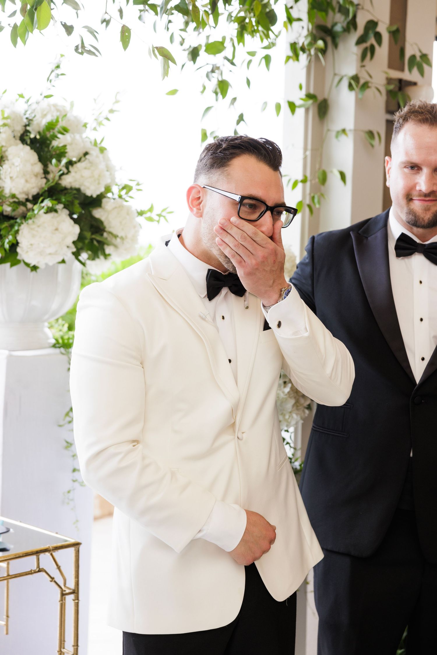 Wedding party member in white tuxedo jacket showing emotional reaction during ceremony.