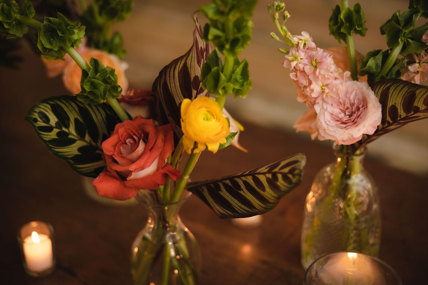 Romantic floral arrangement with roses in clear vases alongside glowing candles in dim lighting.