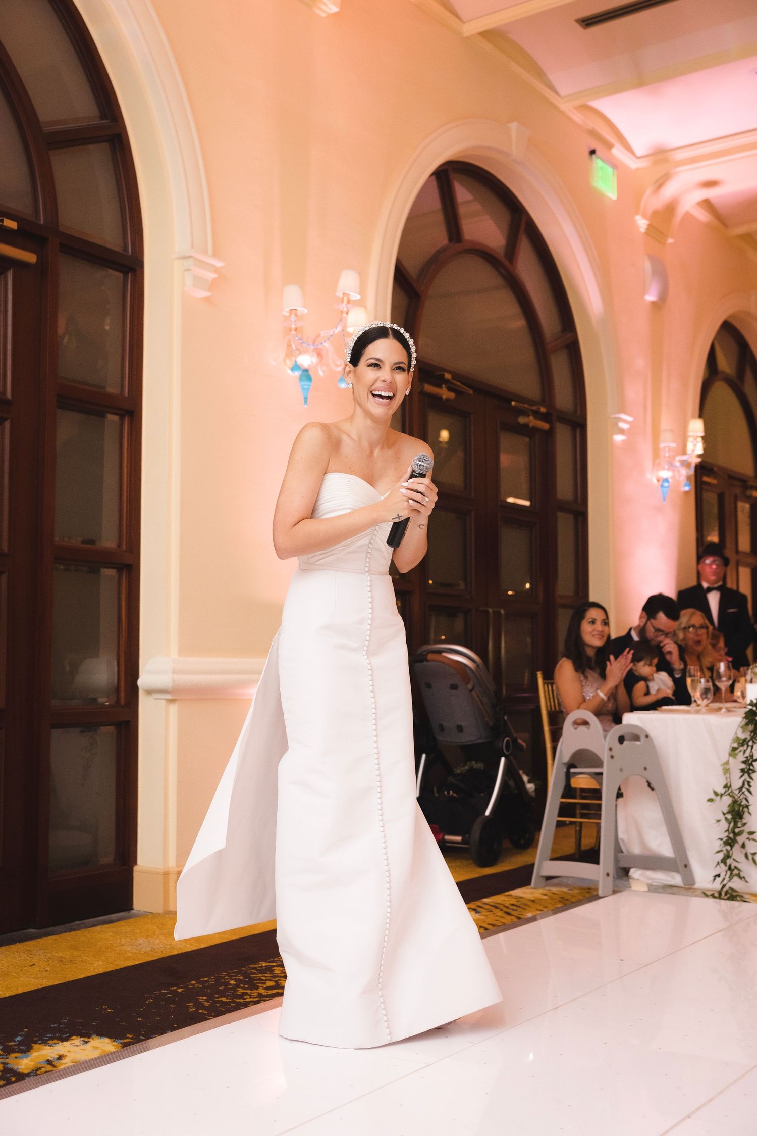 Bride in white strapless gown dancing and celebrating during wedding reception in elegant ballroom with pink lighting.