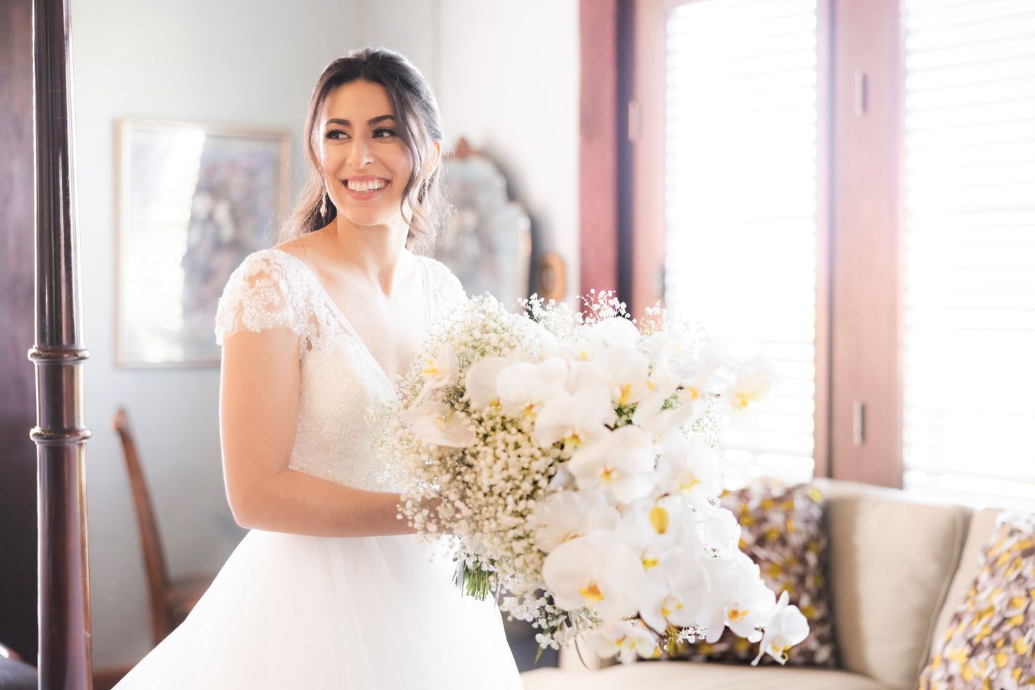 A bride wearing a traditional white lace wedding dress poses with a fresh floral bouquet in a softly lit room.