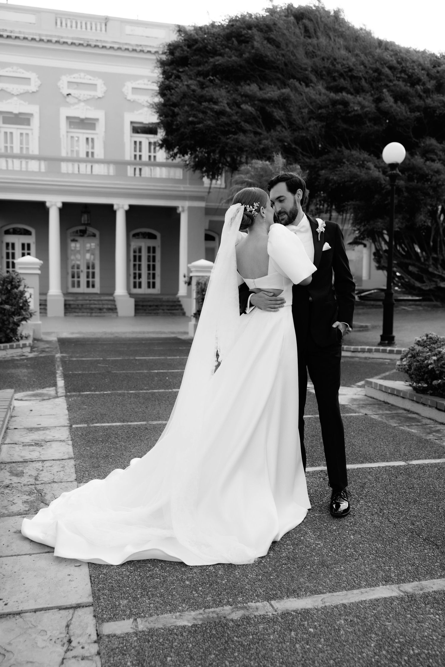 A couple shares an intimate moment in formal wedding attire outside a stately colonial-style building with white columns.