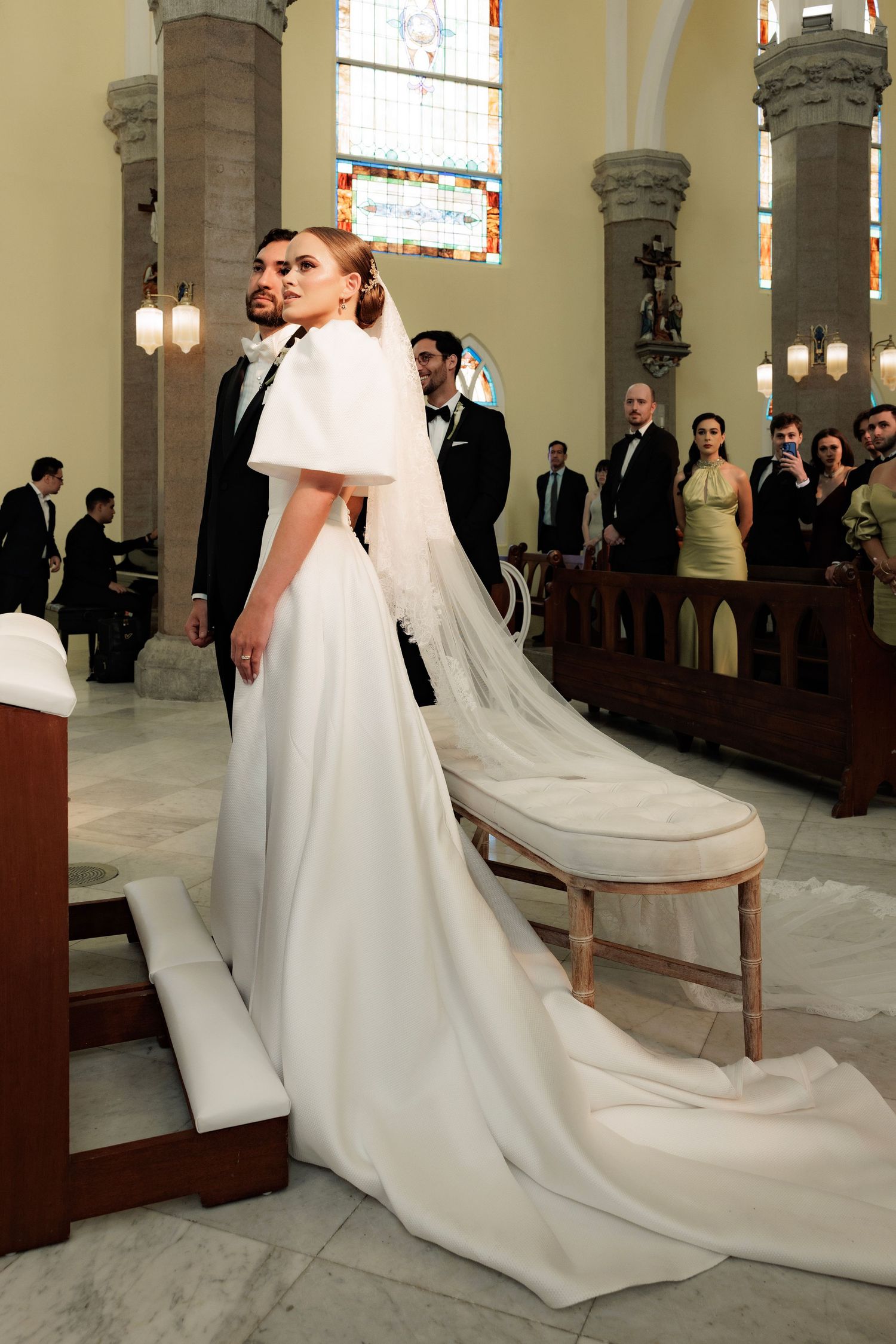 An elegant bride in a white gown with puff sleeves and long train stands in a grand church setting.