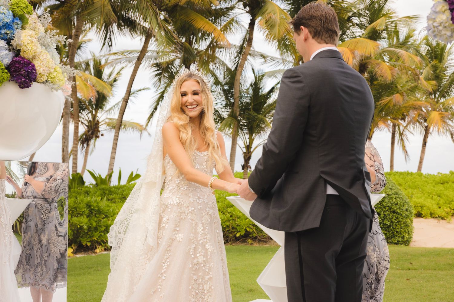 A couple exchanges vows during a beach wedding ceremony surrounded by palm trees and tropical landscaping.