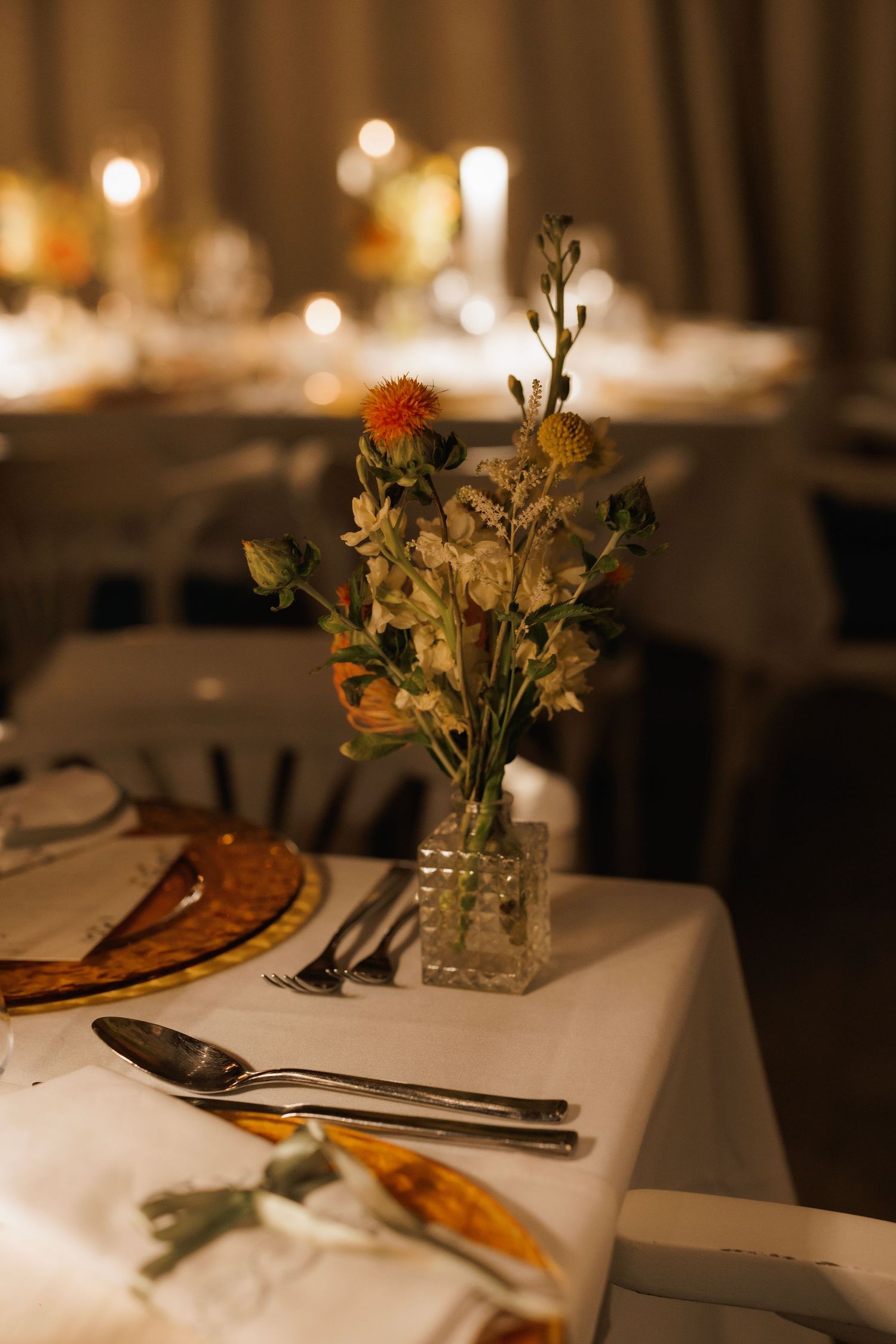 Closeup of an elegant dinner place setting with white tablecloth, warm lighting, and delicate floral centerpieces.
