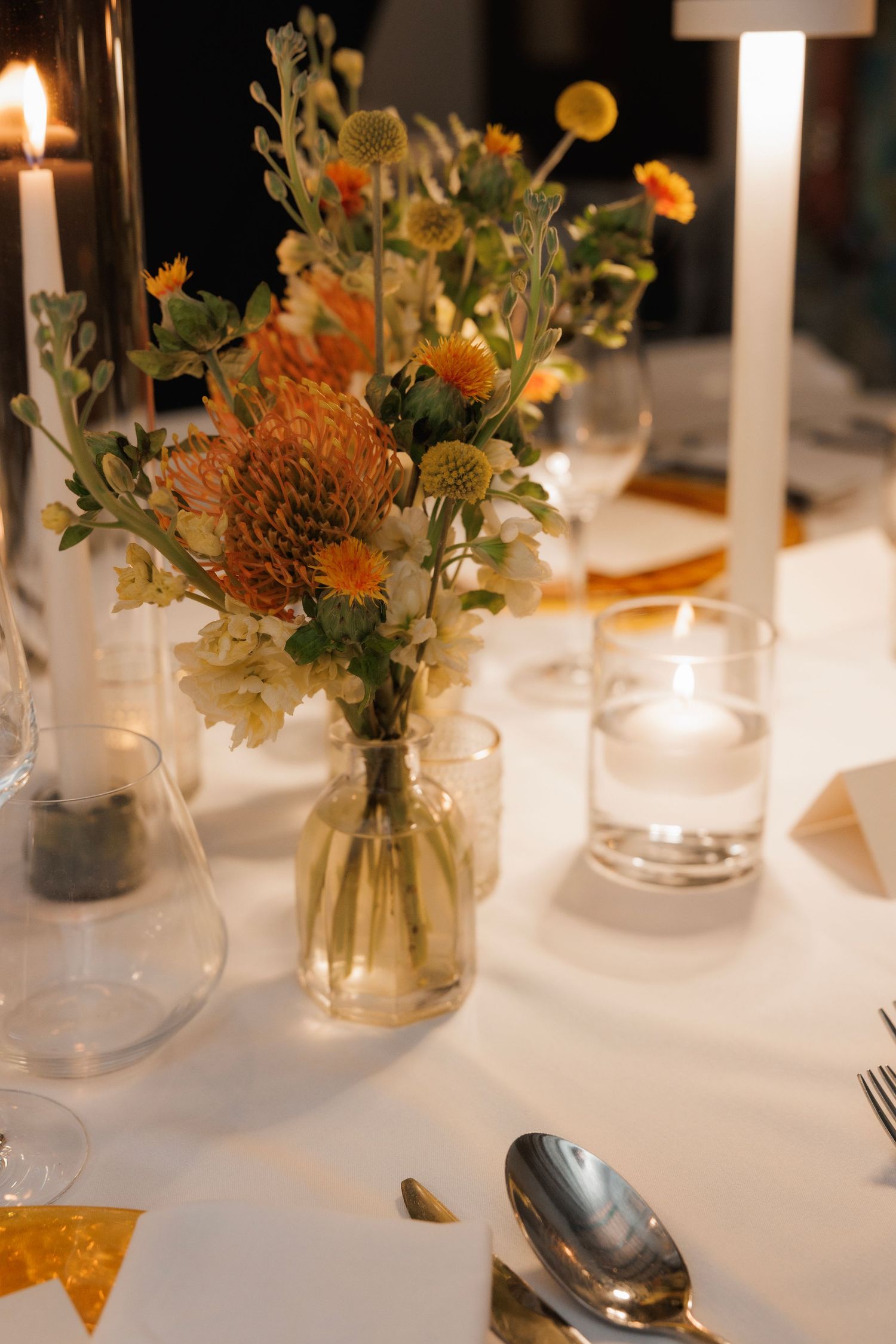 Elegant table setting with orange floral centerpiece in glass vase alongside white candles on crisp white tablecloth.