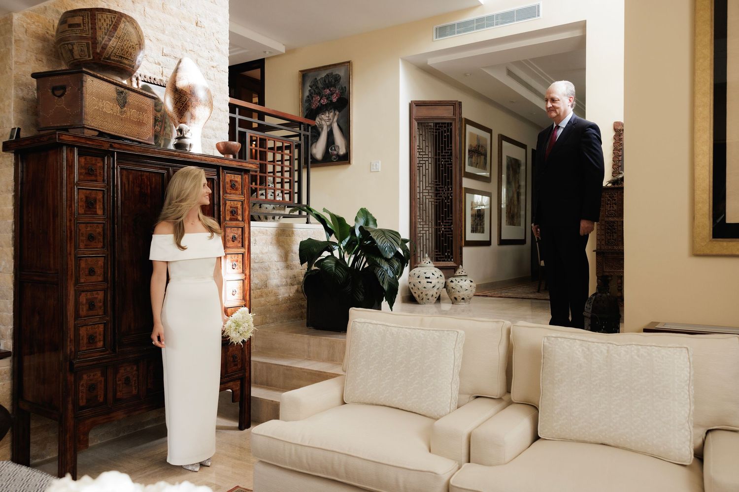Two people stand in an elegant living room with cream-colored furniture and a tall wooden cabinet with antiques.