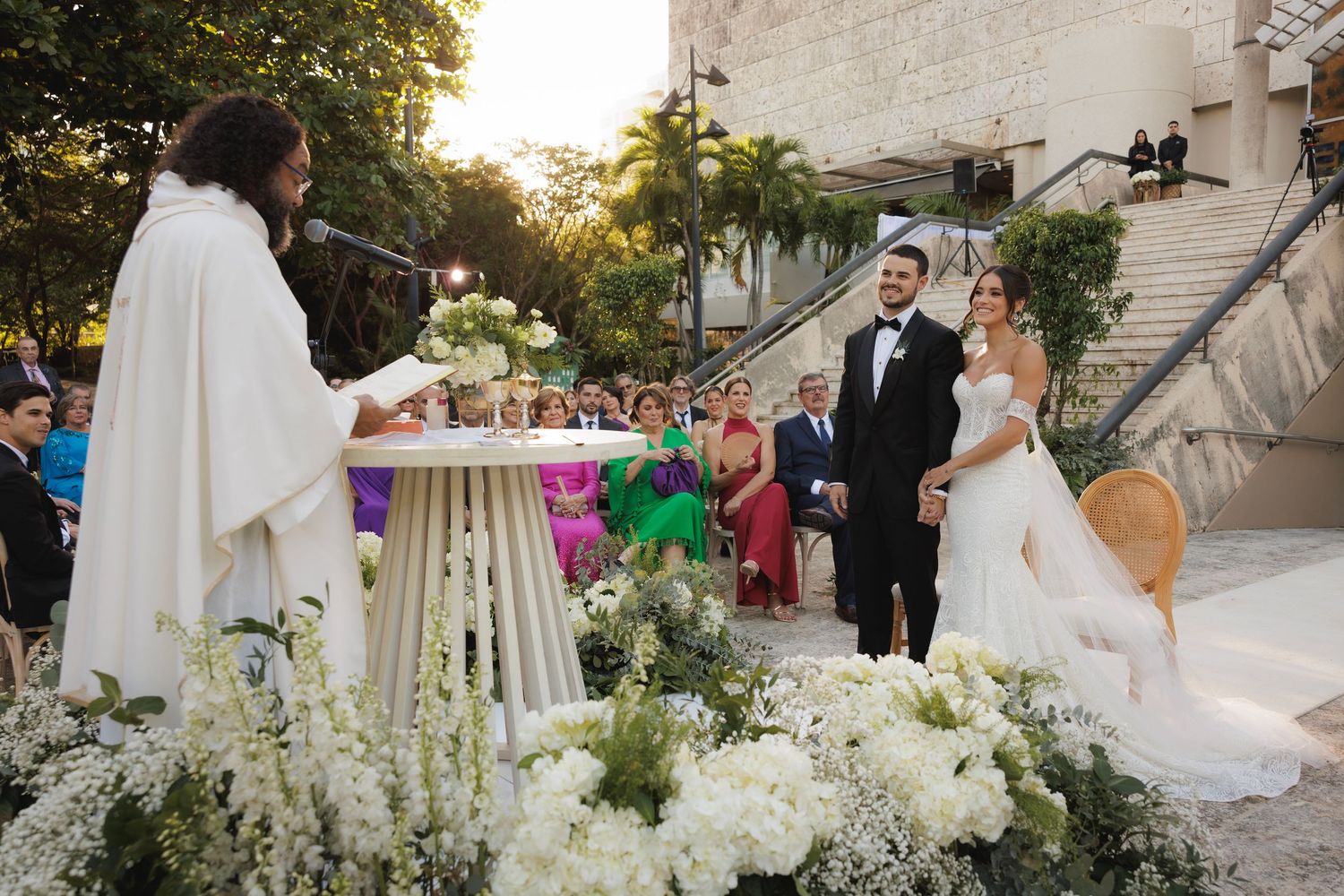 Elegant wedding ceremony setup with white floral arrangements and guests gathered in an outdoor garden setting at sunset.
