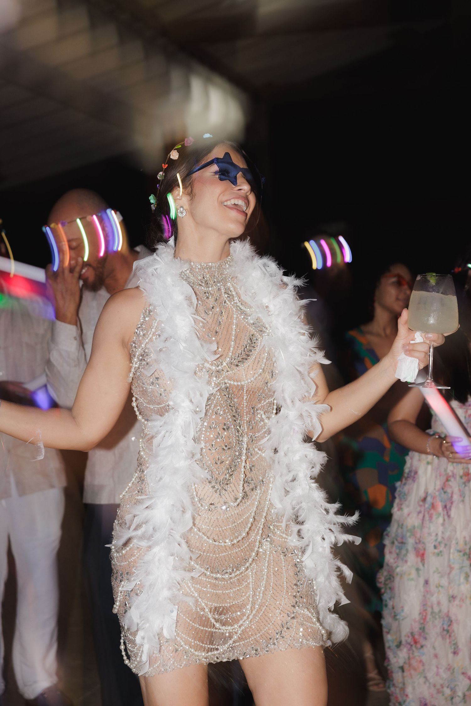 Person dancing in a white feathered costume with pearl details under club lighting at a festive party.