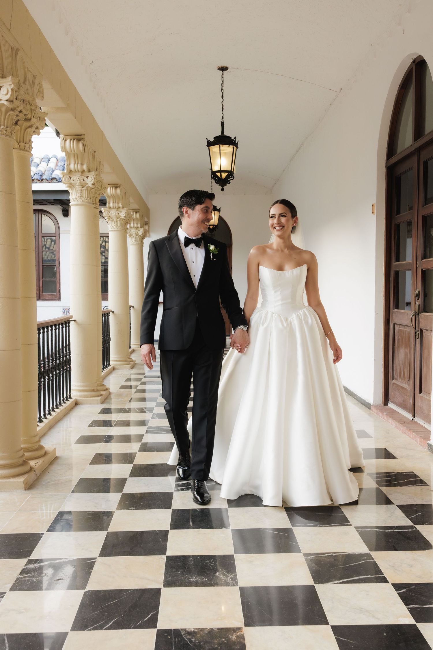 Wedding couple walking playfully along a checkered marble floor in an elegant colonnade.