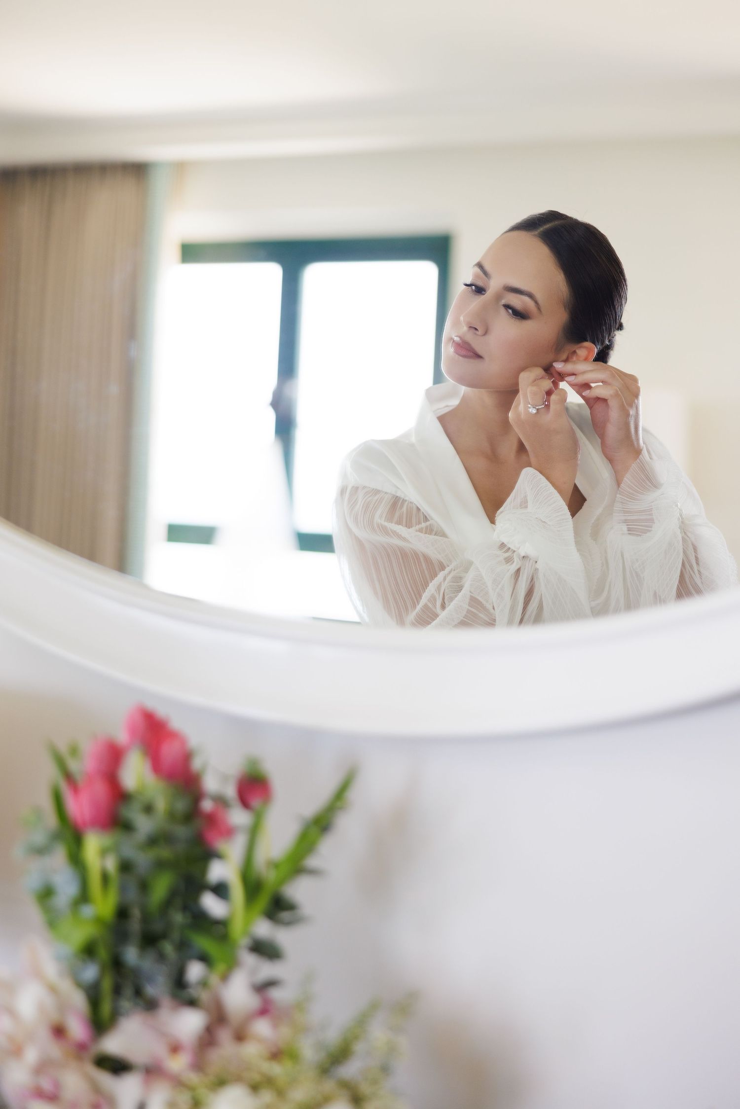 A soft, elegant getting-ready moment with pink flowers and white drapery in the foreground.