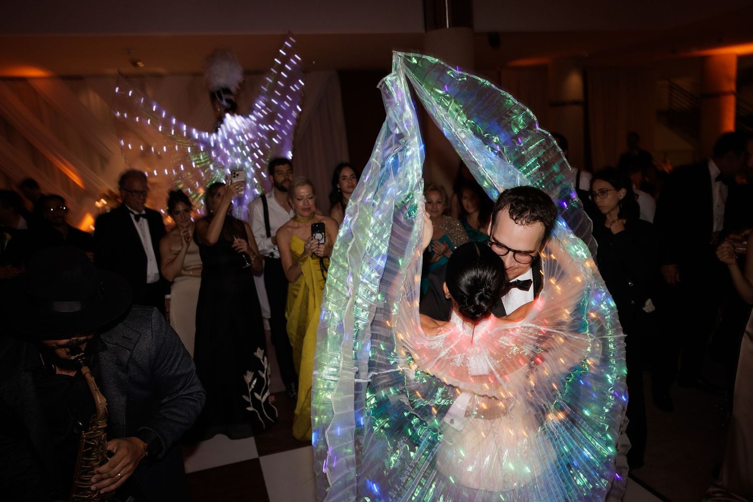 Wedding guests dance and celebrate under colorful LED wing displays at a reception.