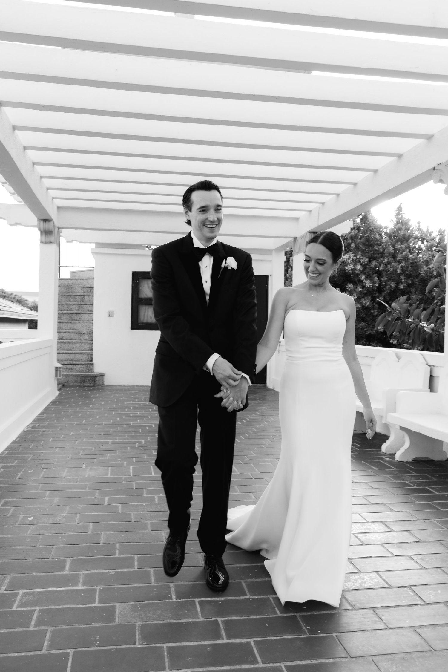 A happy couple in formal wedding attire walks together and laughs under a white pergola on their special day.