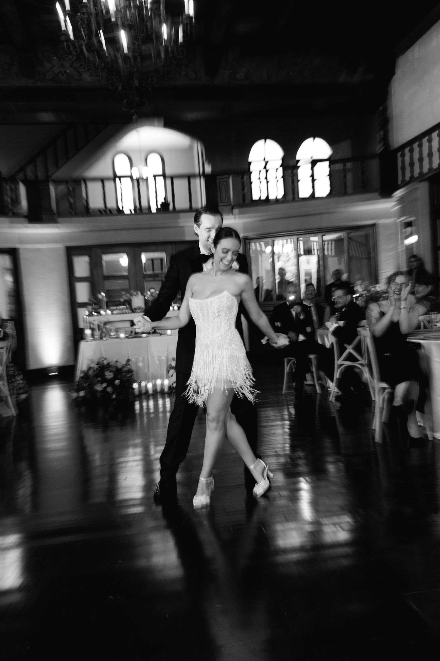 A couple performs an elegant dance together in a grand ballroom with chandeliers and arched windows.